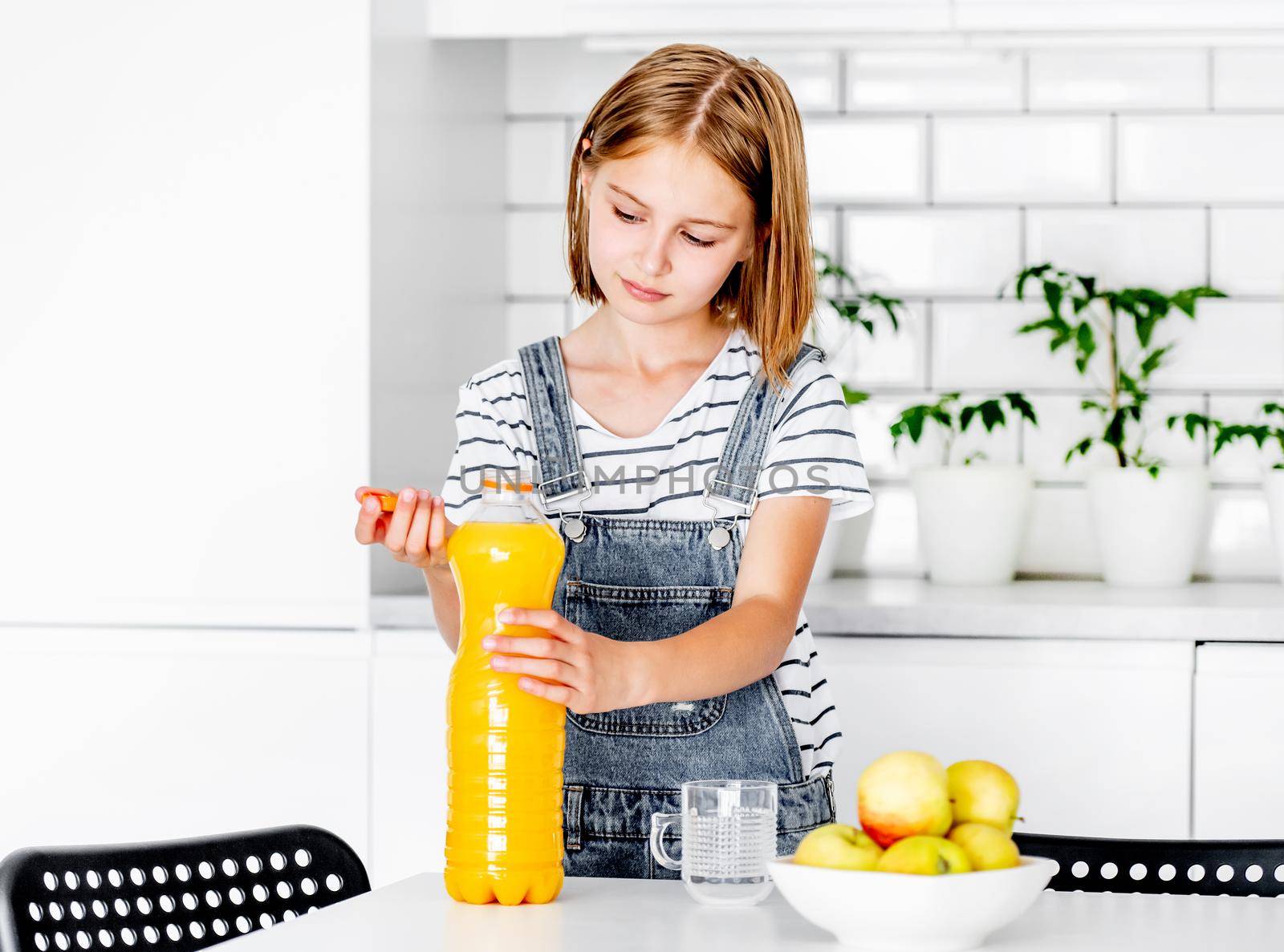 Preteen girl with orange juice bottle and glass at kitchen. Pretty child kid with vitamin fruit drink at home