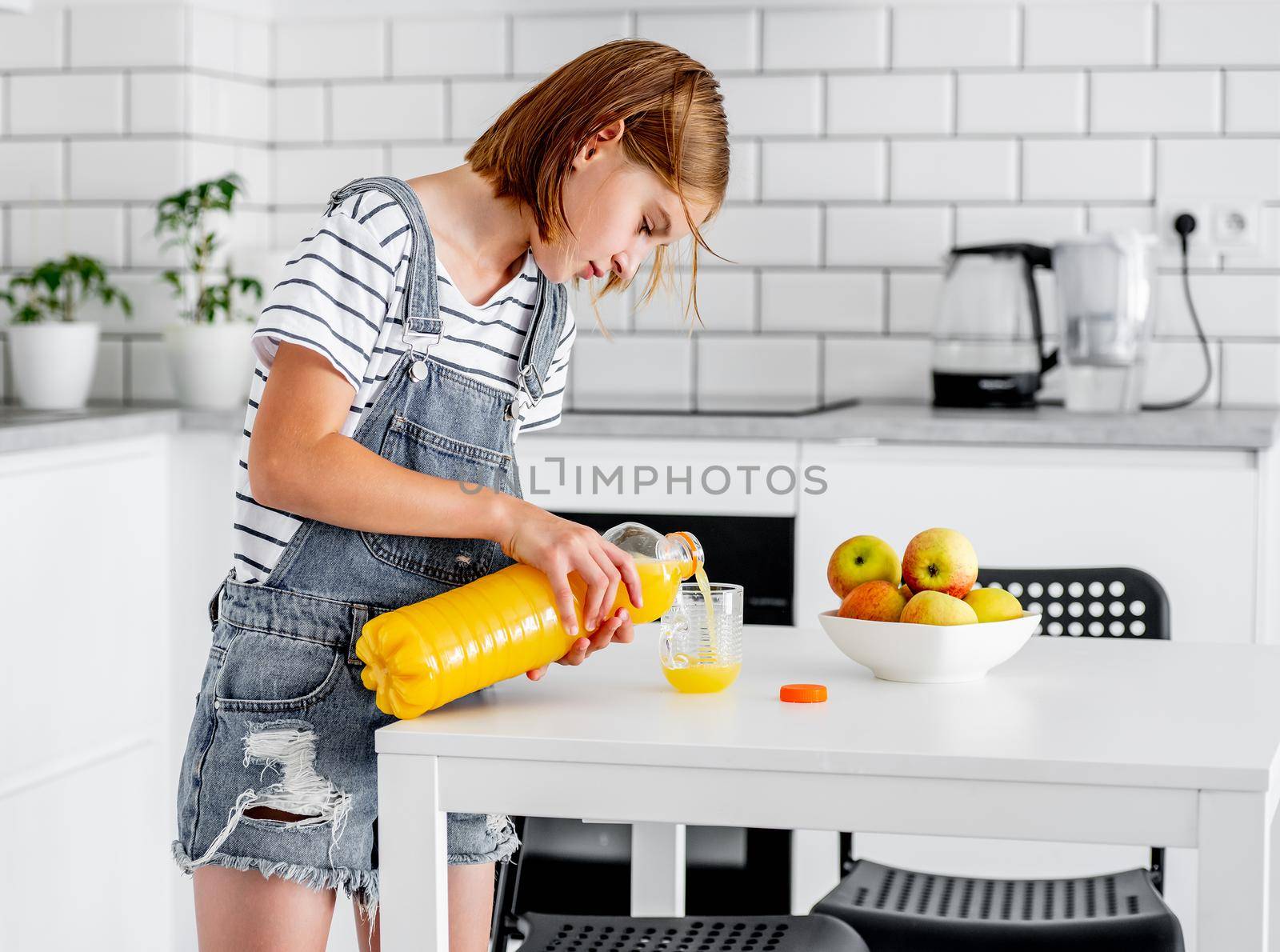 Preteen girl pours orange juice from bottle to glass at kitchen. Pretty child kid with vitamin fruit drink at home