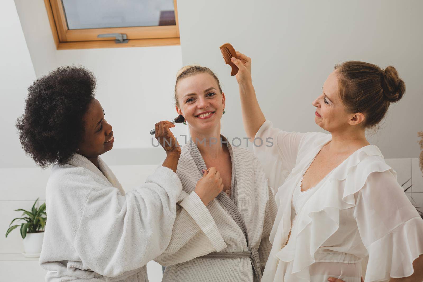 Three smiling young women in bathrobes have fun at the spa and beautify each other