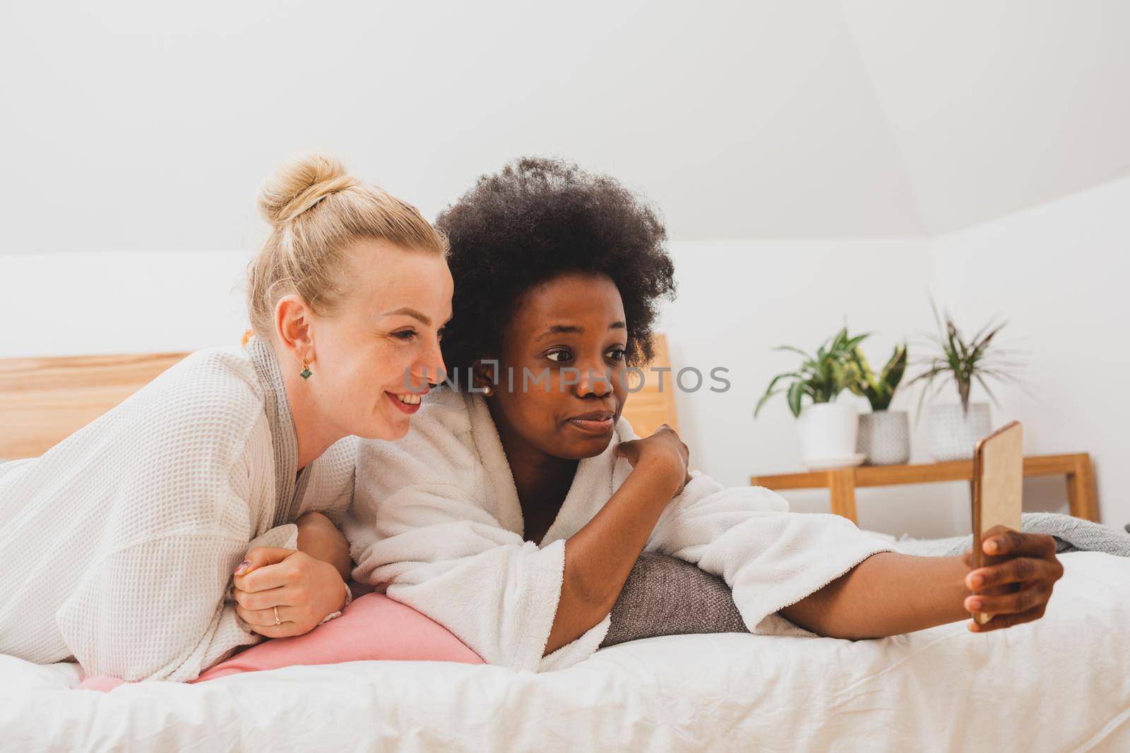 Two young women are lying on the bed and take selfies. The women in bathrobes are resting after spa treatments