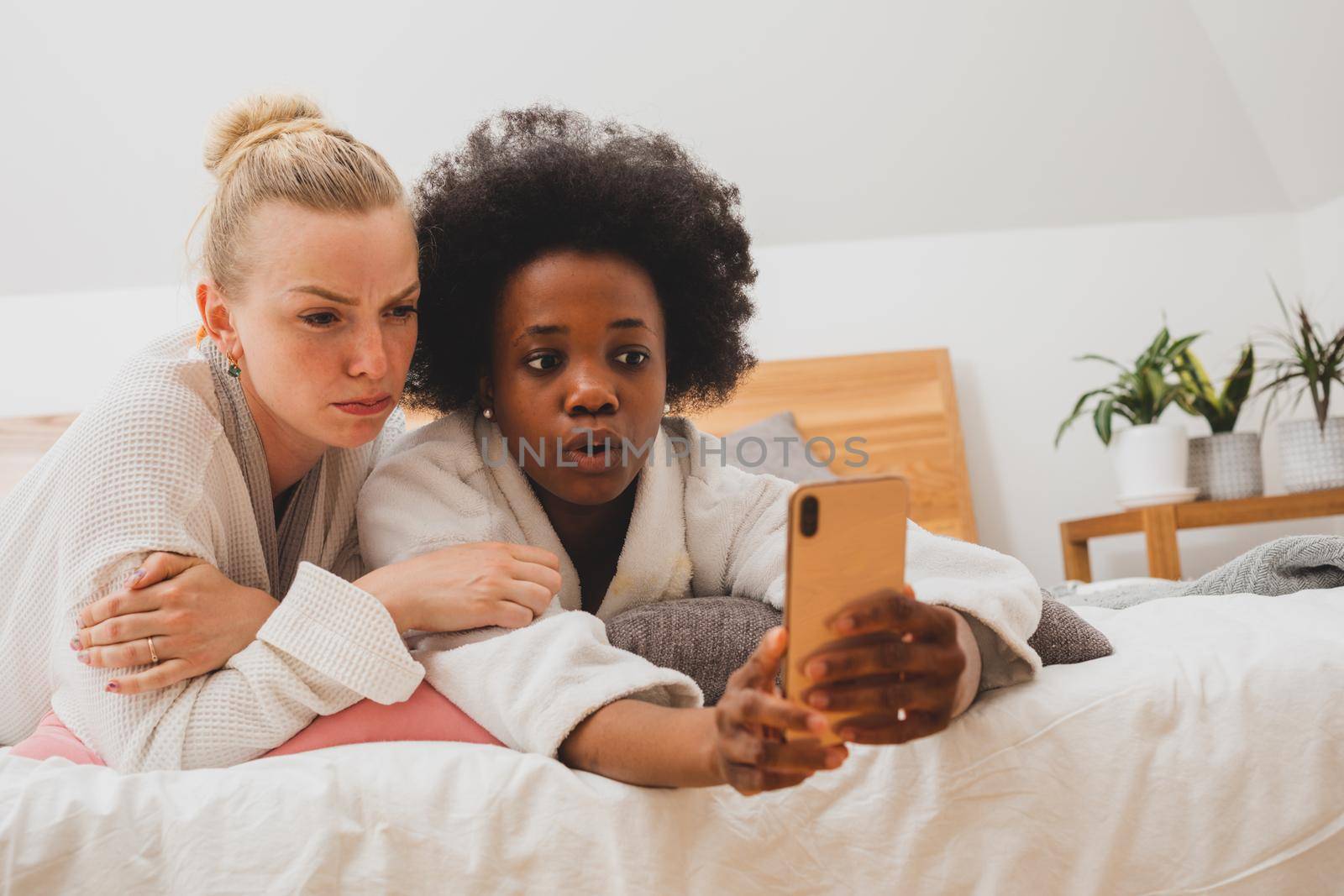 Two young women of different nationalities have a video chat at the spa. The women in bathrobes are lying on the bed and communicating to using a smartphone