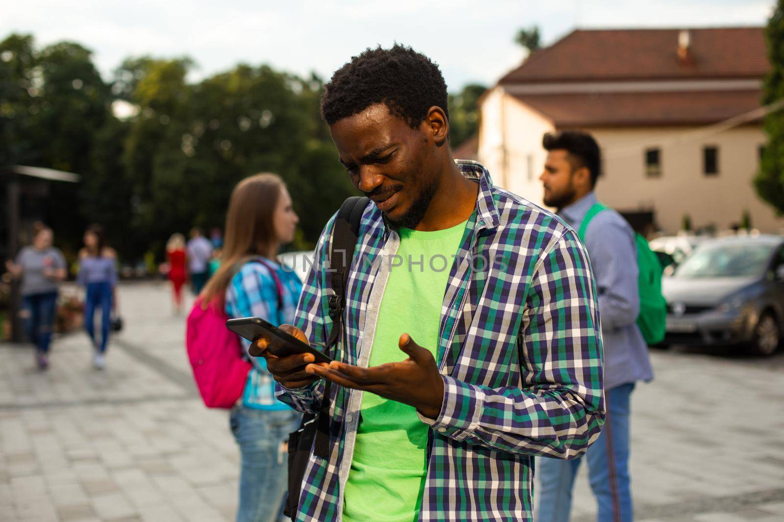 The young african student with a bag outdoors is holding a phone. The man in a shirt is looking for road to home with help navigation