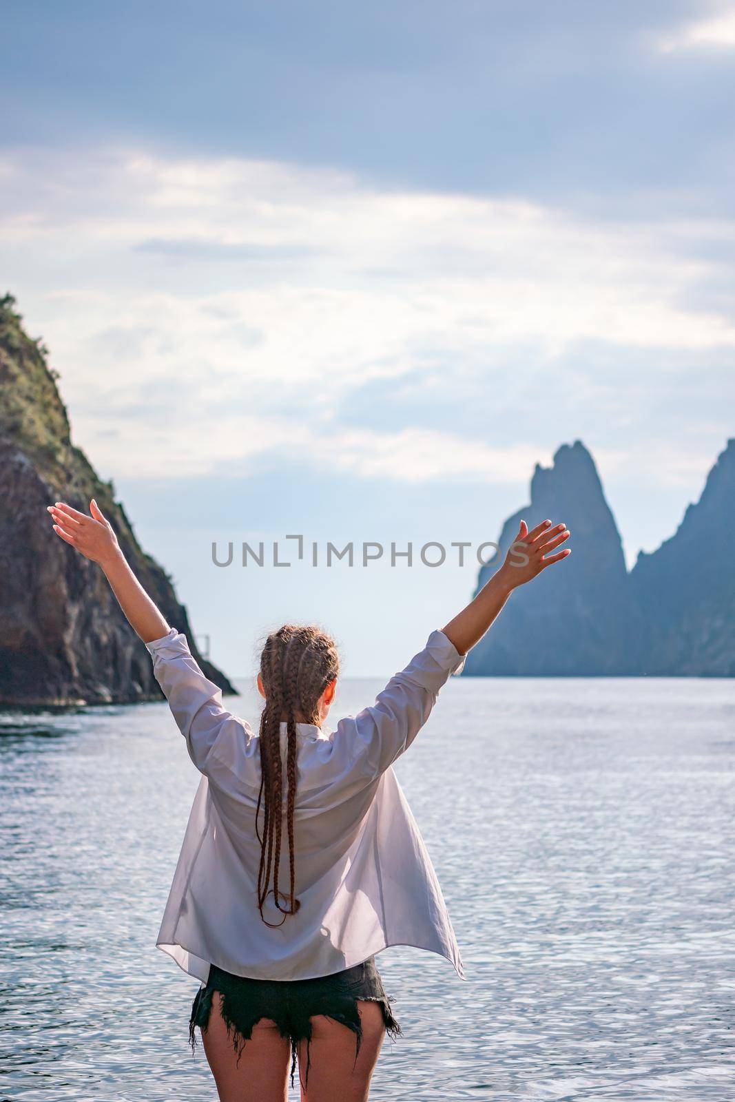 The girl stands on the shore and looks at the sea. Her hands are raised up. She wears a white shirt and her hair is in a braid