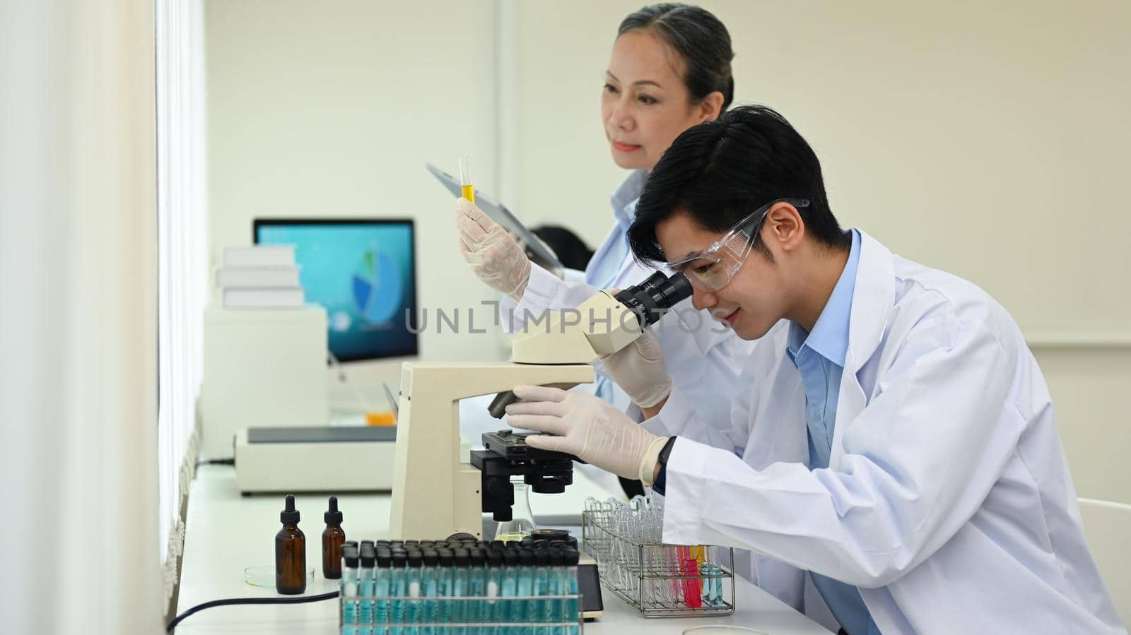 Young male microbiologist in white coat looking through a microscope, examining samples in research laboratory.
