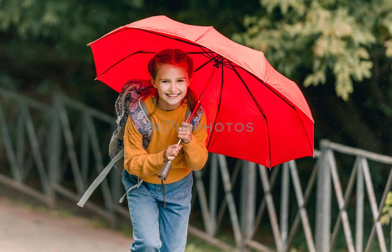 School girl with backpack and umbrella outdoors. Pretty child kid walking in rainy weather