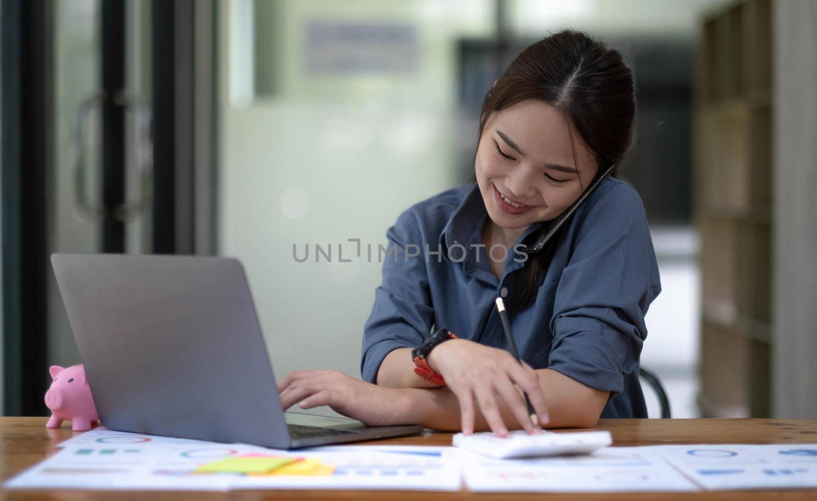 Portrait of Asian young female talking on the phone with a business customer while using a calculator recording data on her work computer. by wichayada