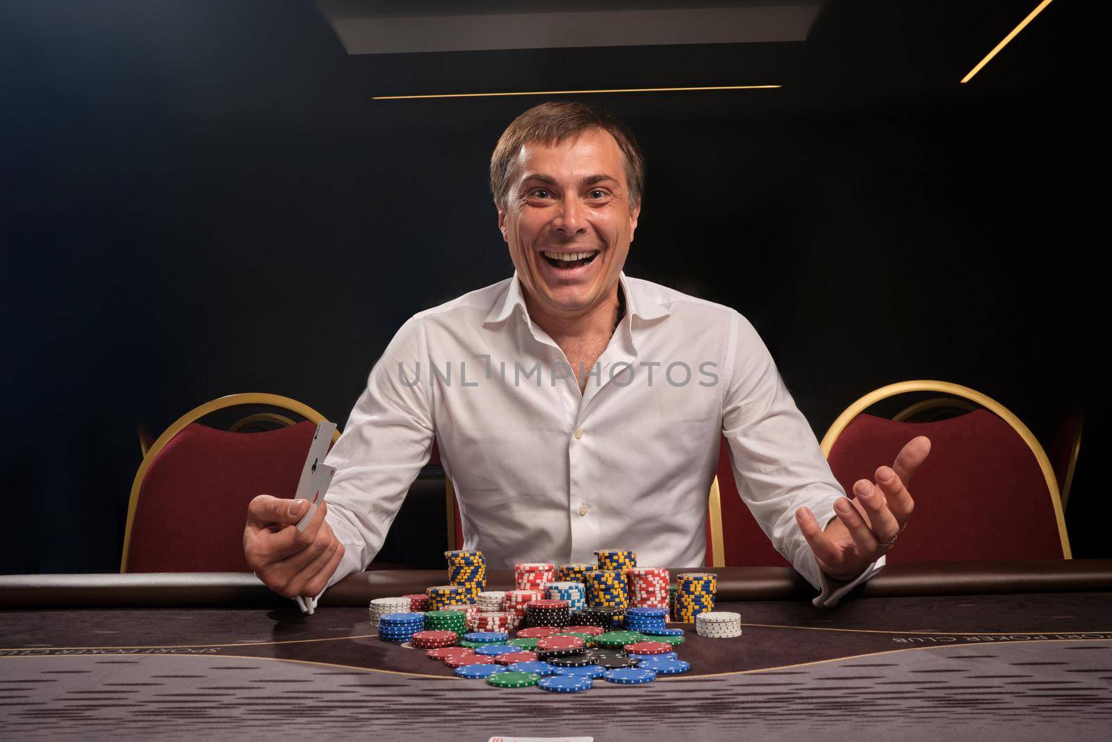 Young smiling man in a white classic shirt is playing poker sitting at the table in casino. He is rejoicing in winning and looking at the camera. Gambling for money. Games of fortune.