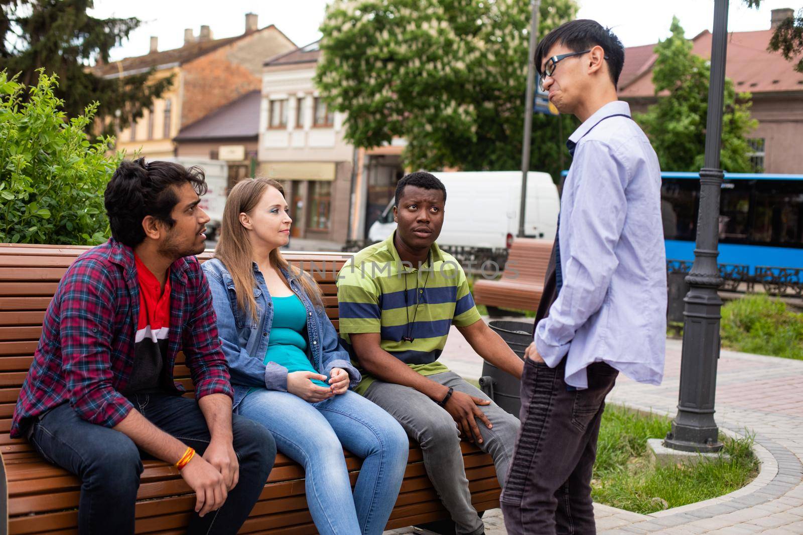 Three handsome multiethnic young men sitting on wooden bench together with lovely blond girl. College students at city park. Asian young man standing opposite group of classmates.