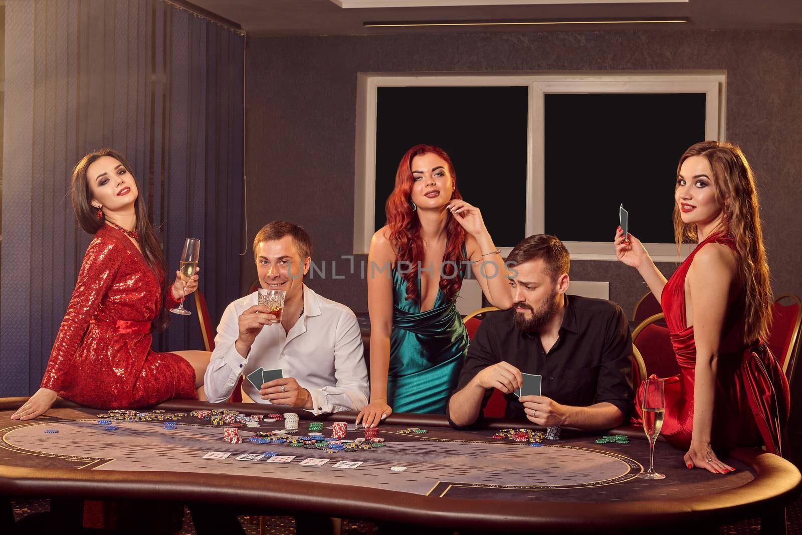 Group of a young players are posing at a poker table in casino and looking at the camera. Youth are making bets waiting for a big win. Gambling for money. Games of fortune.
