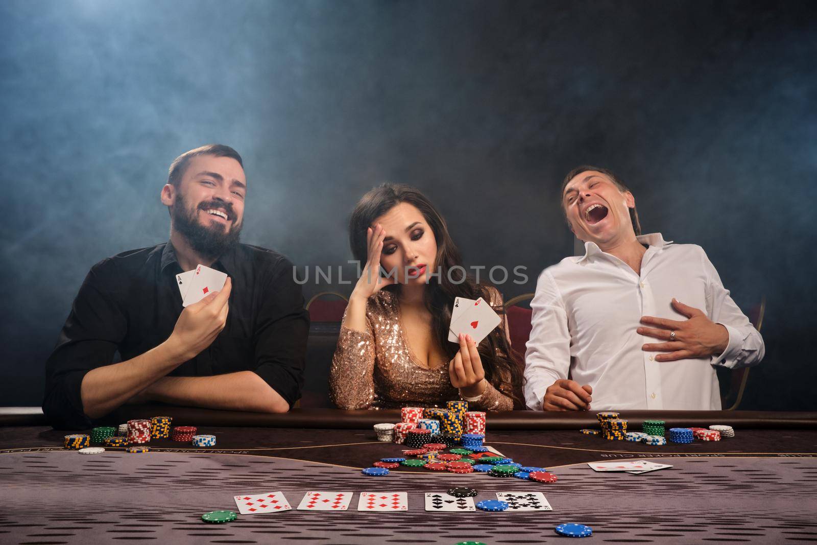 Group of a young friends are playing poker at a casino in smoke. Two men are laughing and a girl is disappointed. Youth are making bets waiting for a big win. Gambling for money. Games of fortune.