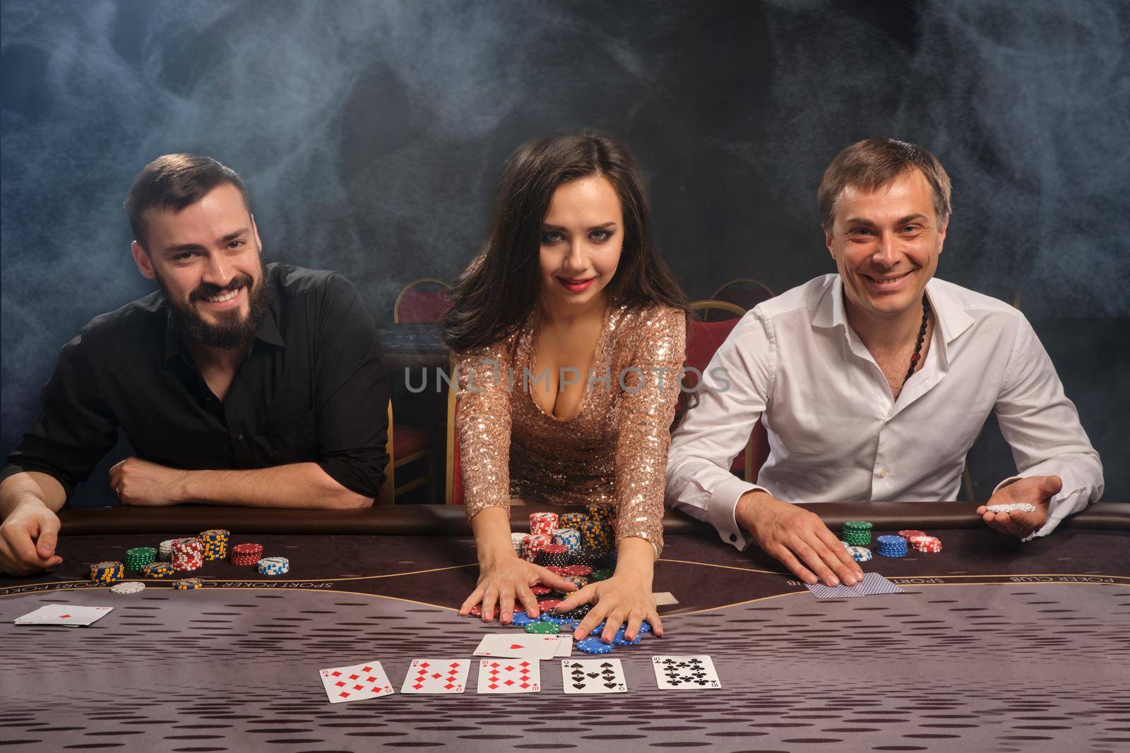 Young cheerful friends are playing poker at casino in smoke and looking at the camera and smiling. Youth are making bets waiting for a huge win. Gambling for money. Games of fortune.
