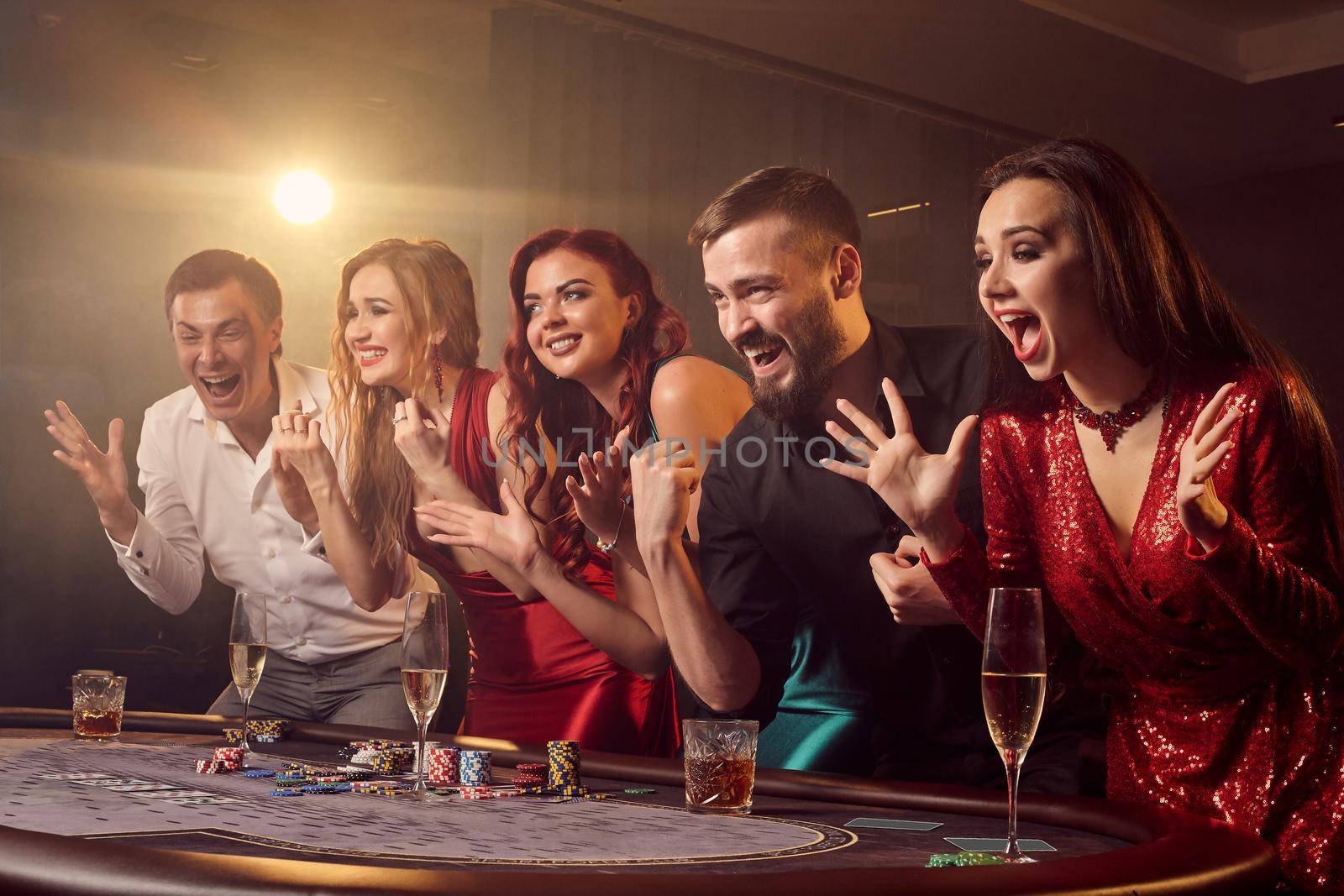 Group of a young joyful friends are rejoicing in their poker victory at casino in a ray of a spotlight. Youth are making bets waiting for a big win. Gambling for money. Games of fortune.