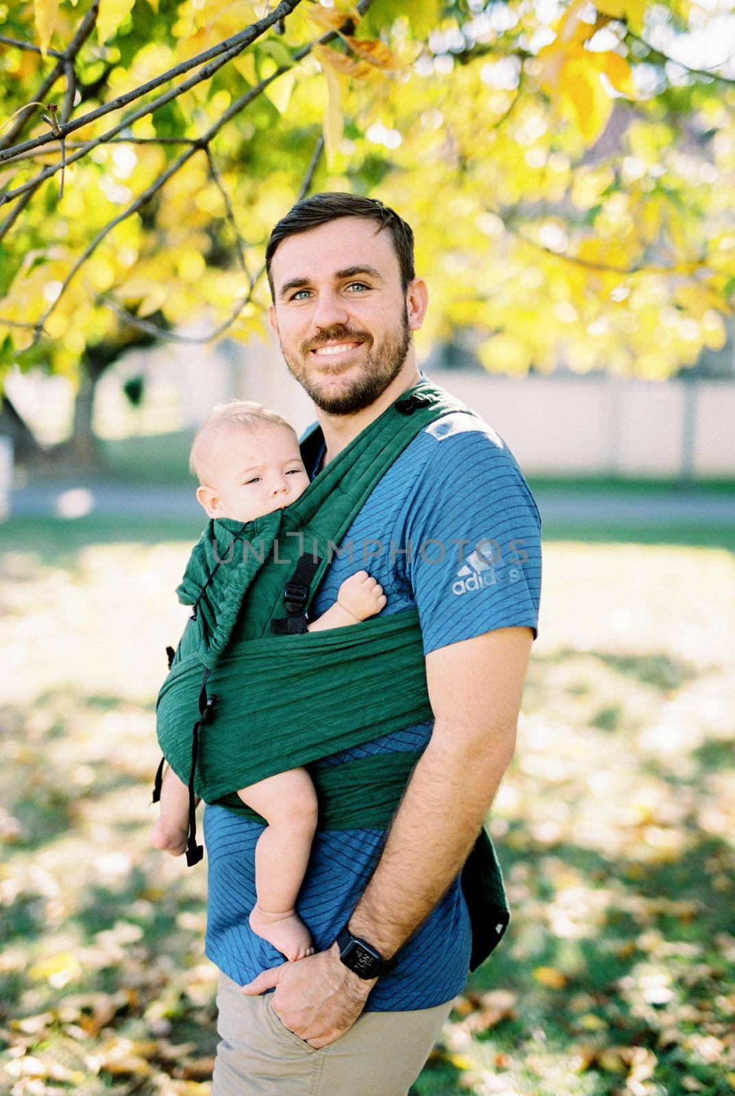 Dad with a baby in a sling standing under a tree. High quality photo