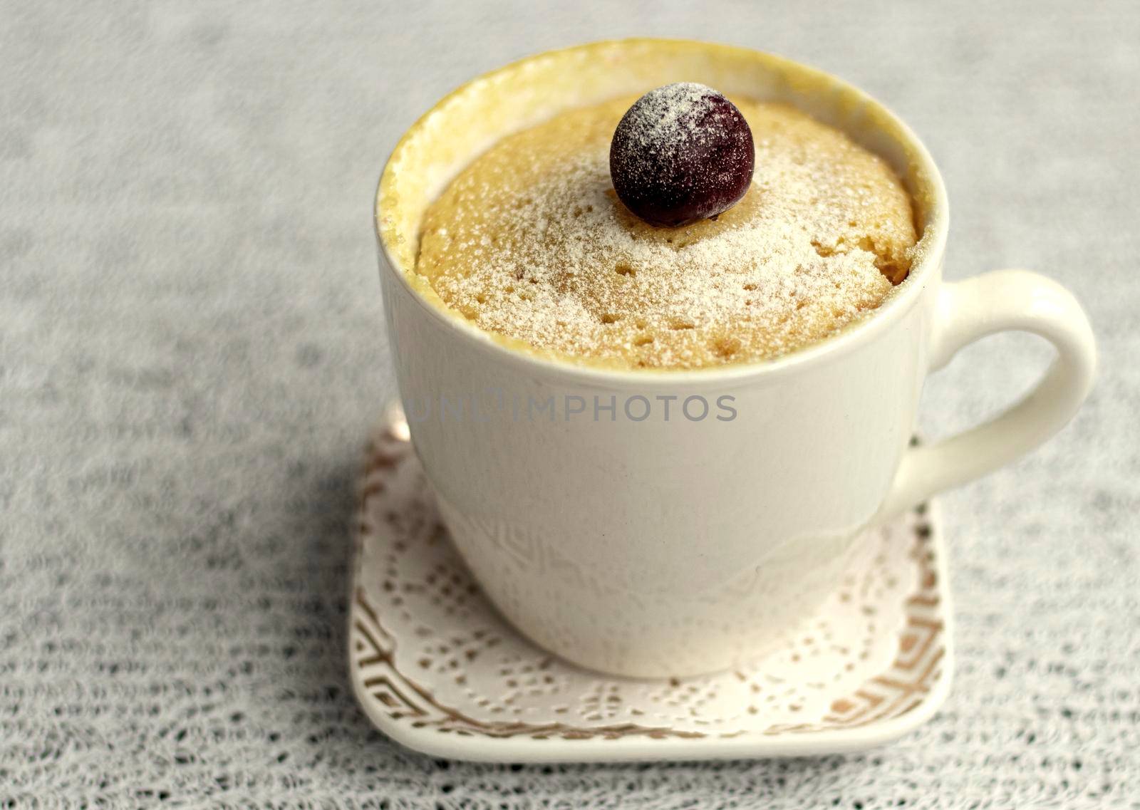 muffin in a mug decorated with cherries. a cup of white flour cake, sprinkled with powder, stands on a saucer, on a light background. Dessert in a mug. Microwave sweet pastries. High quality photo