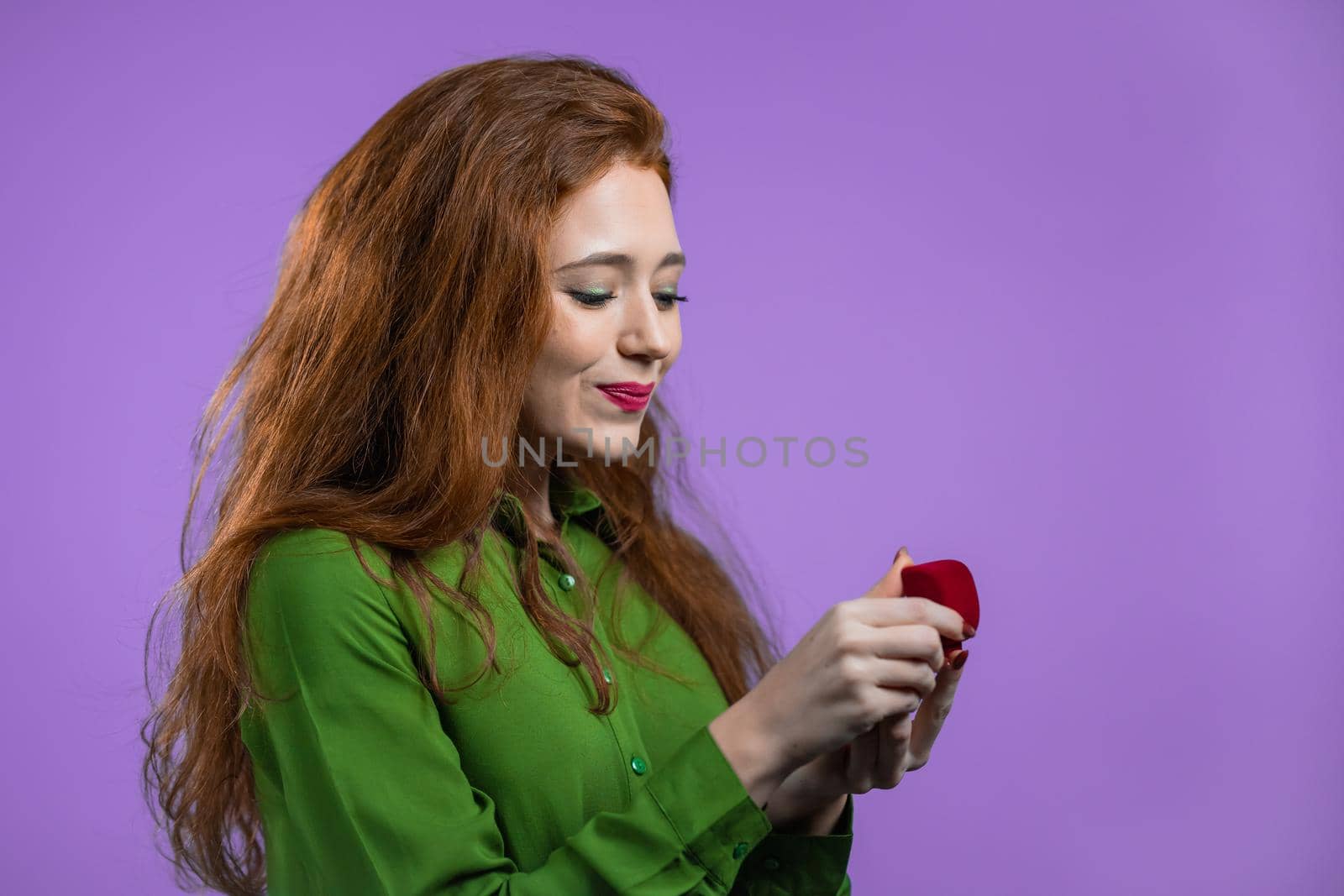 Attractive woman holding small jewelry box with proposal diamond ring on violet wall background. Lady smiling, she is happy to get present, proposition for marriage. High quality photo