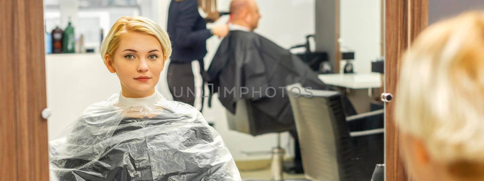Young female client waiting for the hairdresser looking in the mirror in a hair salon, selective focus