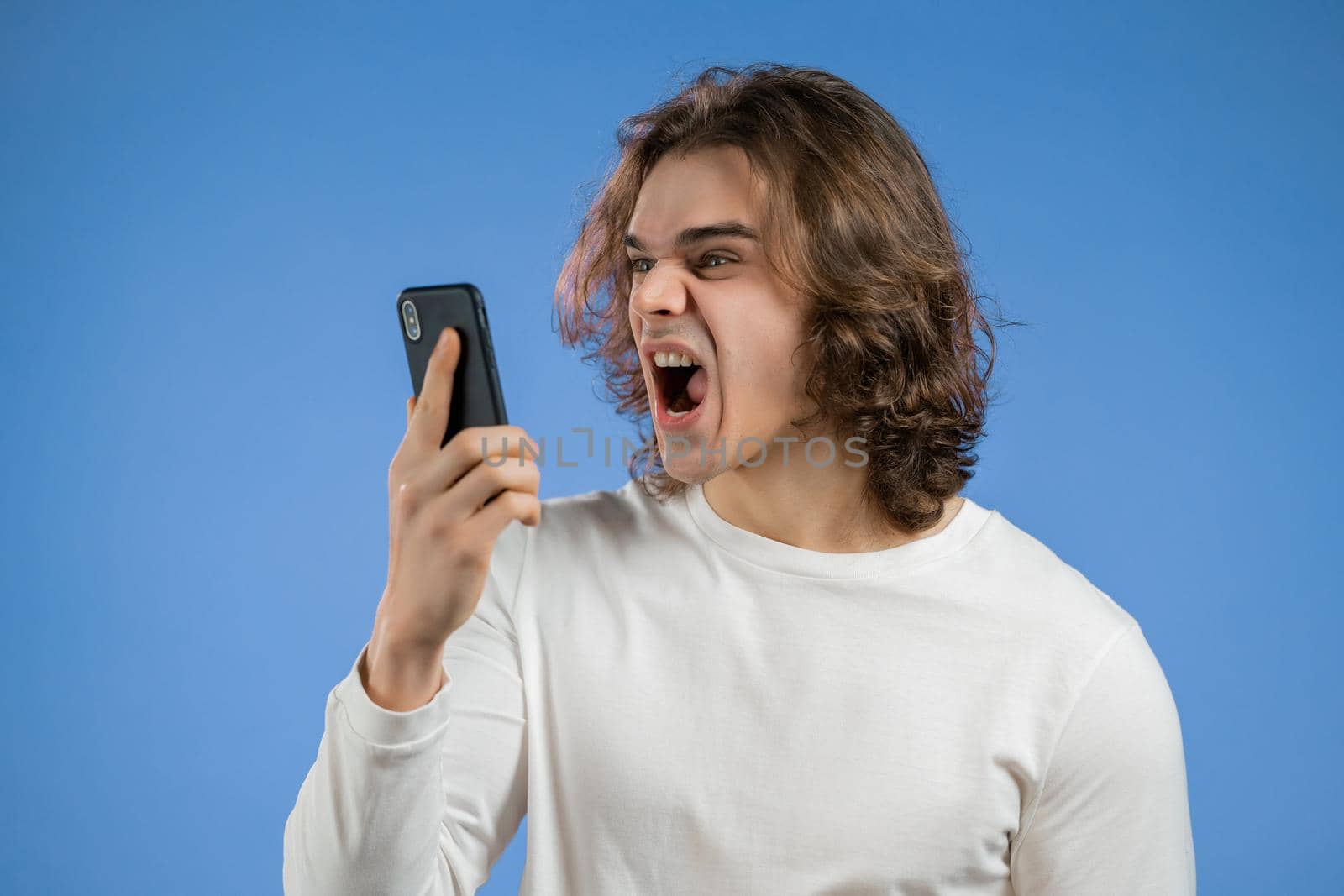 Angry man in white t-shirt screaming down his mobile phone. Stressed and depressed guy on blue background. High quality photo