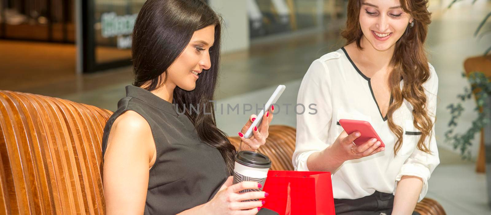 Two young caucasian women looking in smartphones smiling and sitting on the bench in a shopping mall