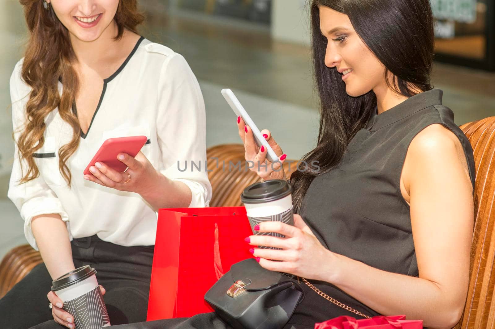 Two young caucasian women looking in smartphones smiling and sitting on the bench in a shopping mall