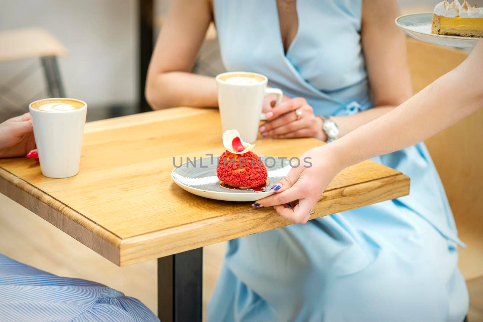 The waiter's hand puts on the table a plate with a red cupcake on the background of a female client in a cafe
