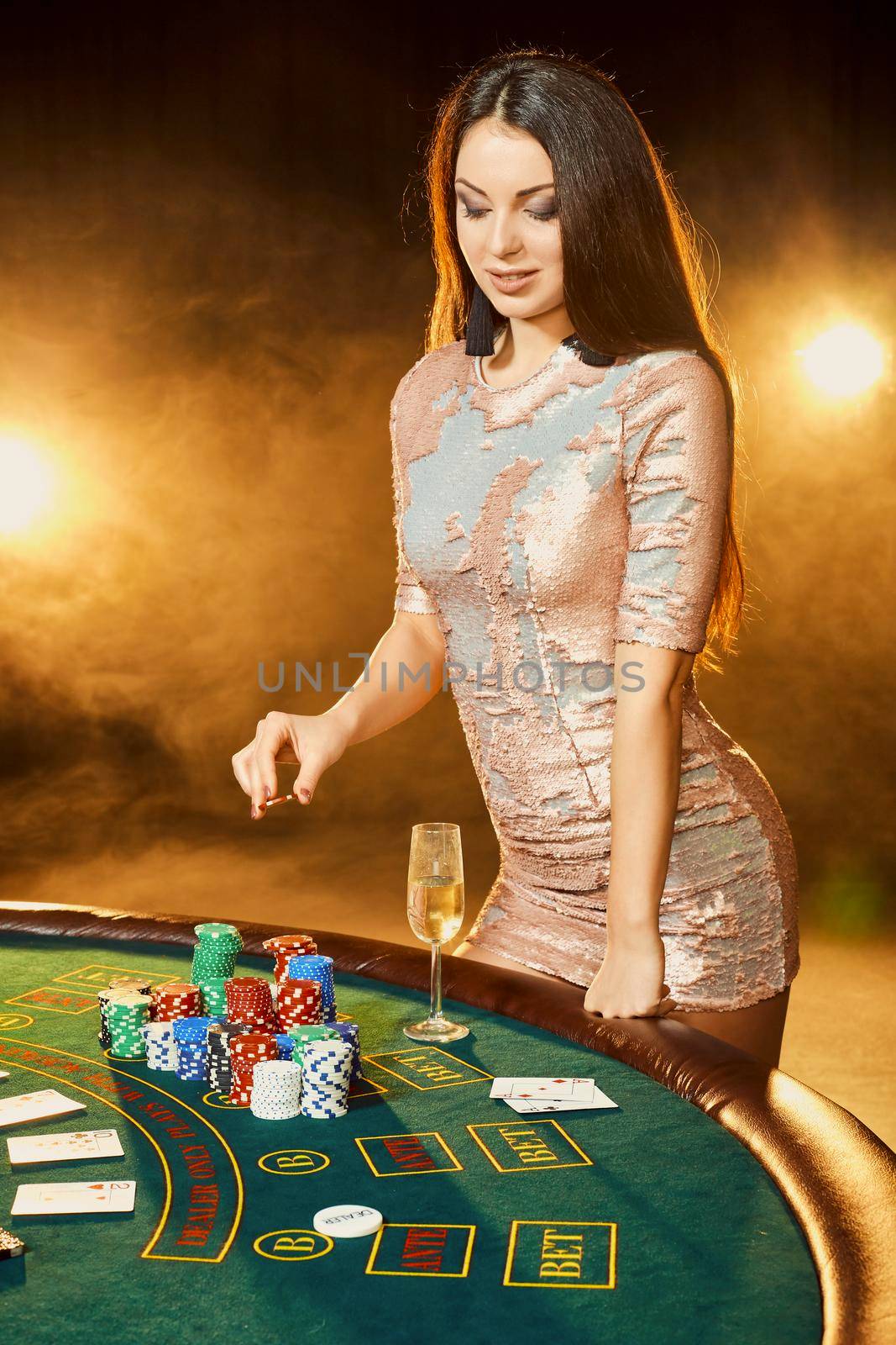 Gorgeous young woman in evening dress with chips in hand standing near poker table with glass of champagne