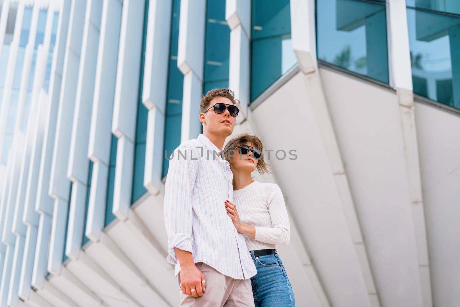 Smiling beautiful woman and her handsome boyfriend. Woman in casual summer jeans. Happy cheerful couple in sunglasses walking business district. Couple posing on the street modern building background
