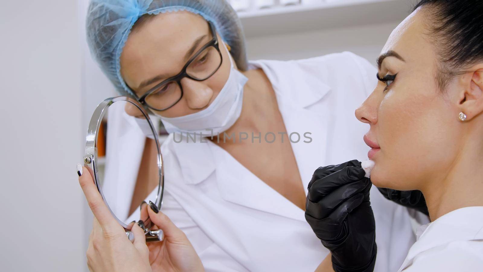 medical office, beautiful woman looking in the mirror, doctor wipes patient lips with a sterile napkin, before injections of hyaluronic acid into lips, cleans the surface with an antiseptic. High quality photo
