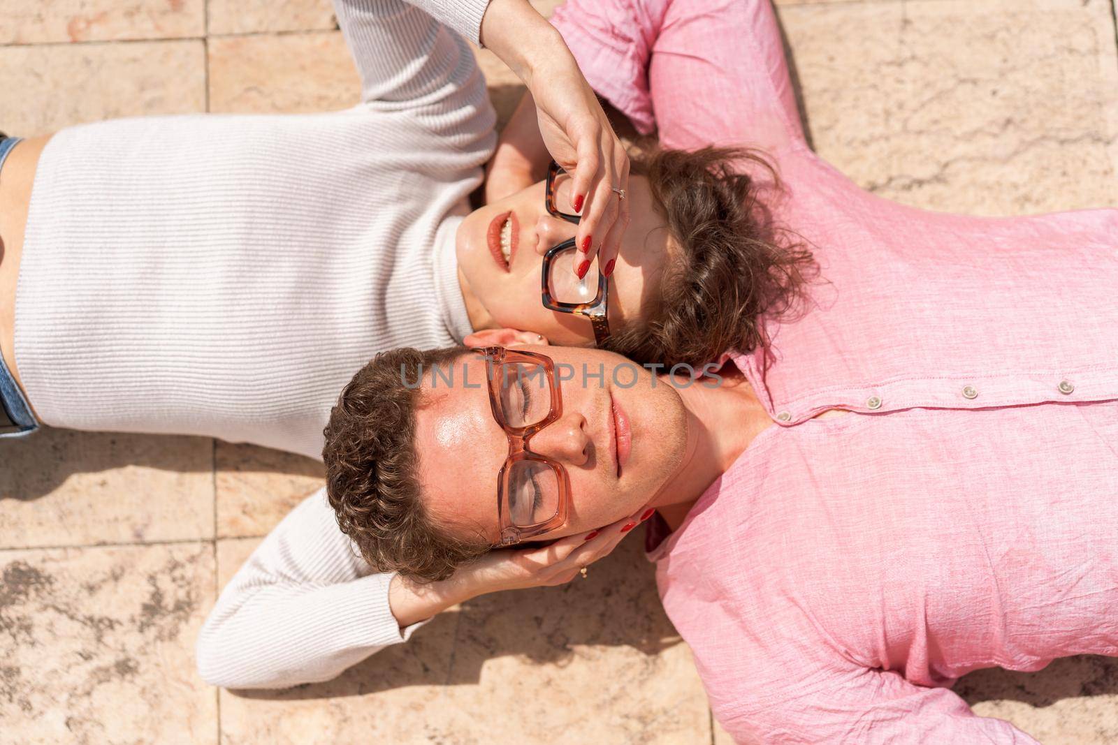 Portrait of two attractive students man and woman wearing stylish glasses lying on floor outdoor and smiling, head to head cheek to cheek, break relax between lesson, work, projects Eyeglasses concept