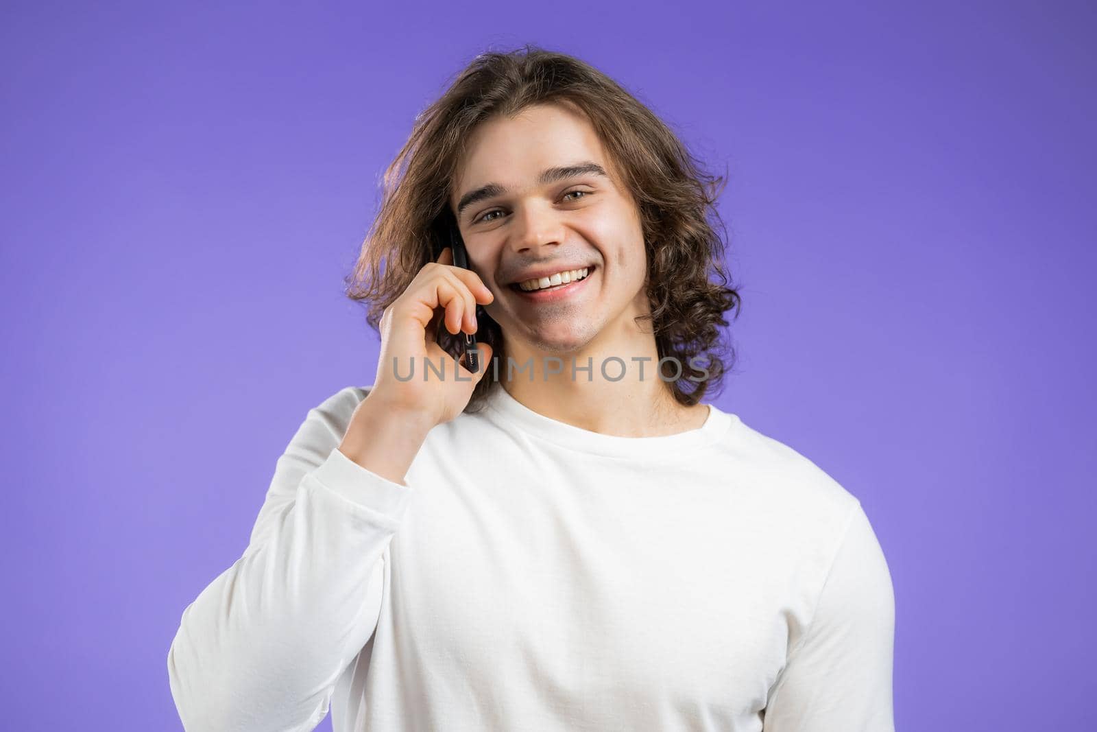 Young smiling man speaks on phone. Guy holding and using smart phone. Violet studio background. by kristina_kokhanova