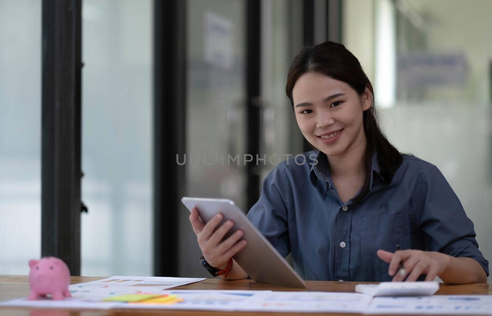 Smiling Asian businesswoman holding a tablet and used calculator at the office. Looking at the camera. by wichayada