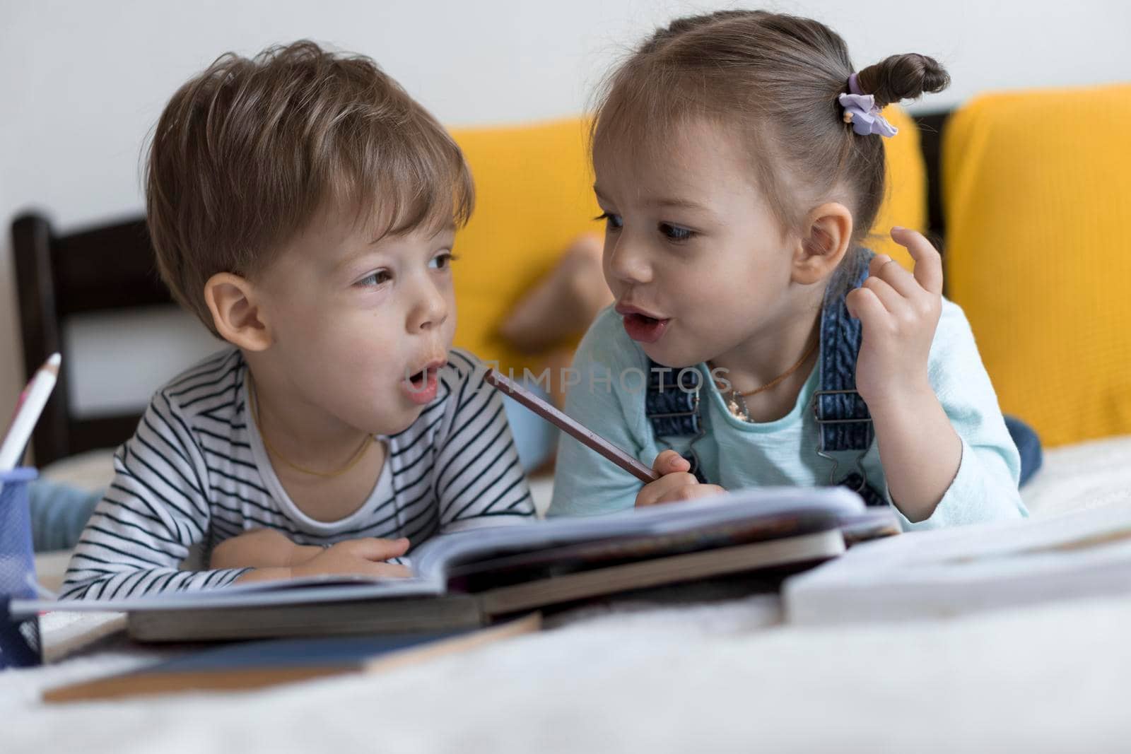 smart smiling preshool toddler children boy and girl draw with pencils lying on their stomach at yellow bed. little cute reader have fun, happy kid on quarantine at home. Friendship, family, education.