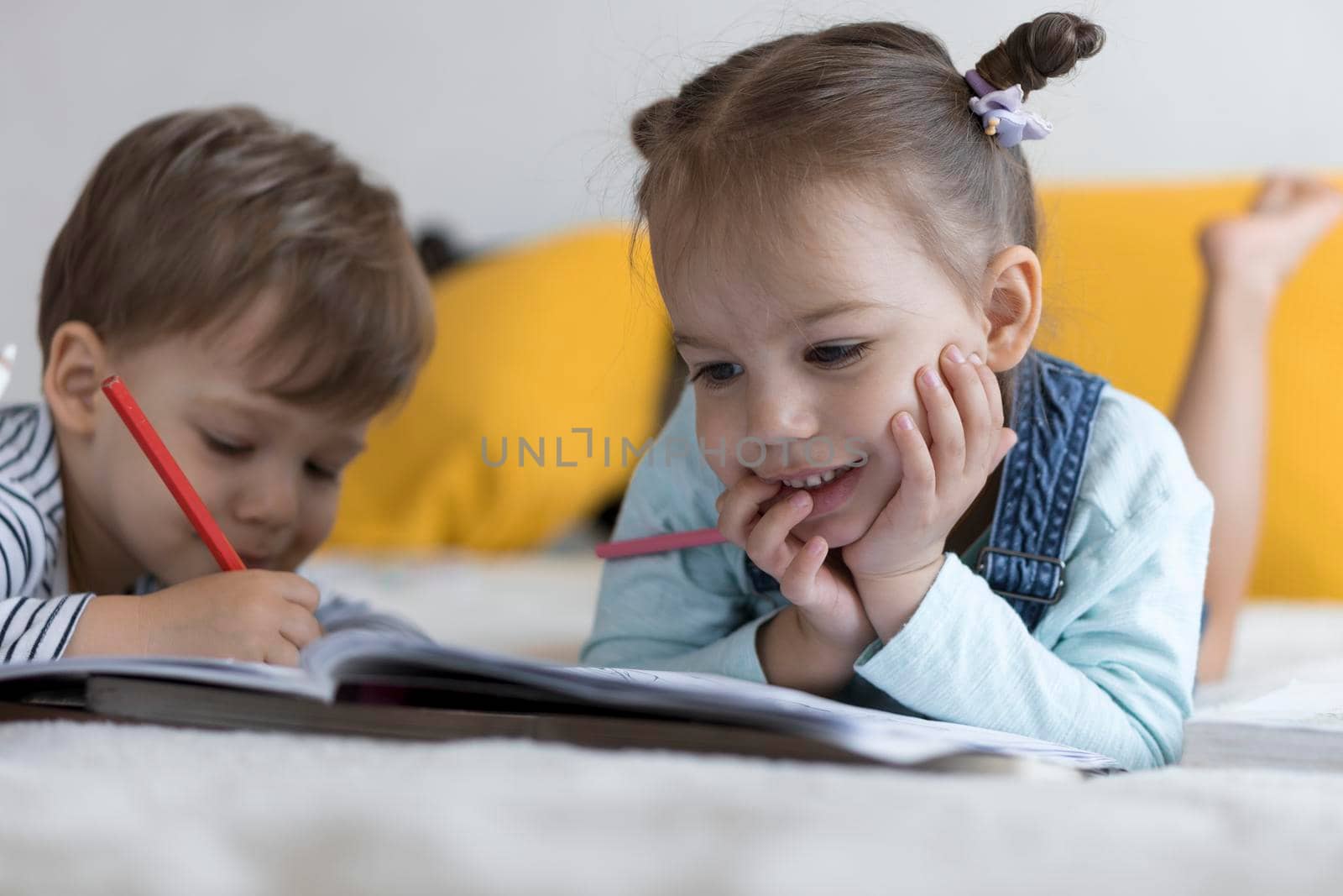 smart smiling preshool toddler children boy and girl draw with pencils lying on their stomach at yellow bed. little cute reader have fun, happy kid on quarantine at home. Friendship, family, education.