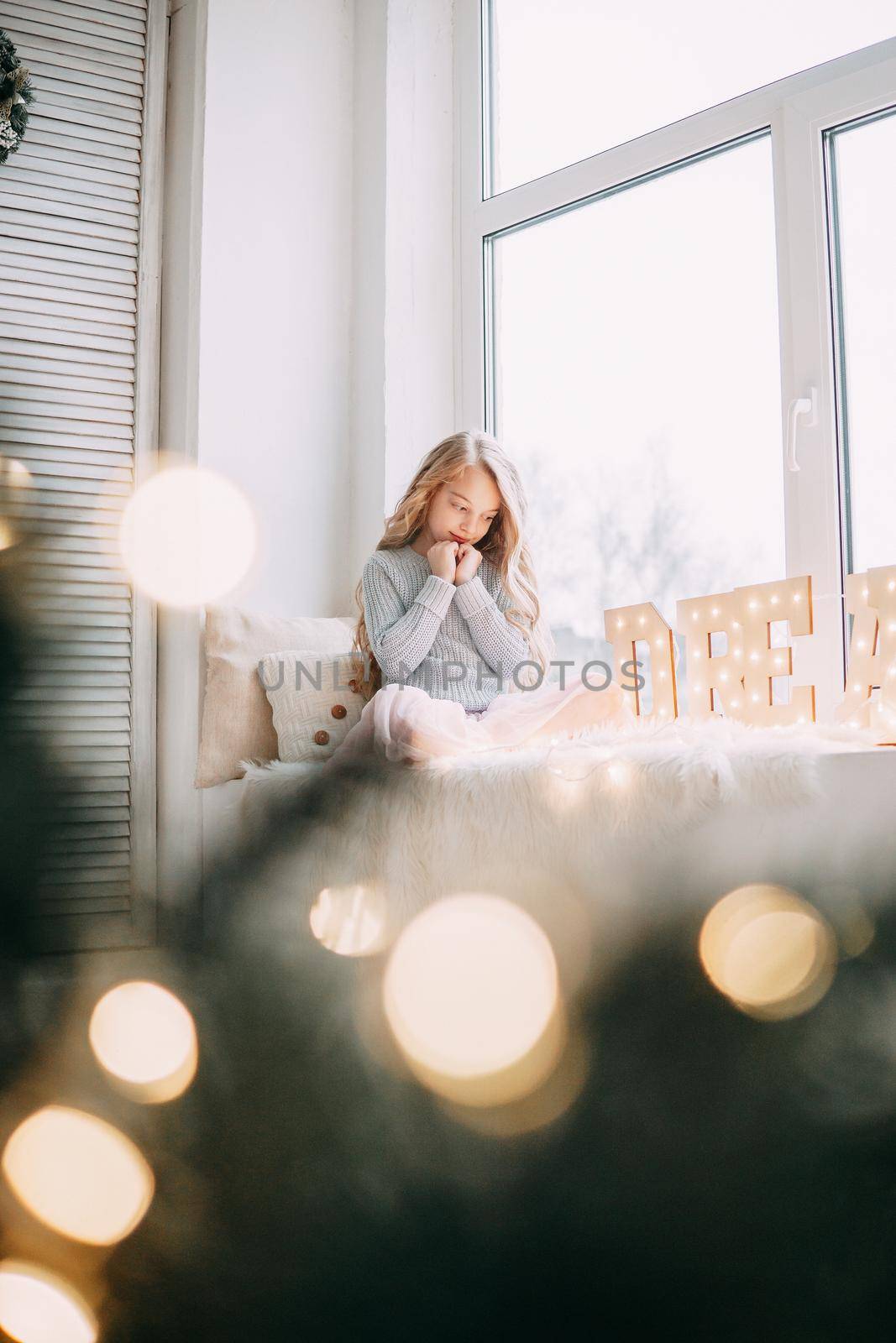 A beautiful blonde girl is sitting on the windowsill with a gray rabbit in a Christmas atmosphere. The concept of pet care.