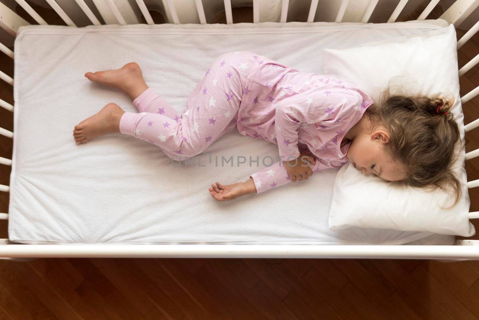 top view Cute little 3-4 years preschool baby girl kid sleeping sweetly in white crib during lunch rest time in pink pajama with pillow at home. Childhood, leisure, comfort, medicine, health concept.