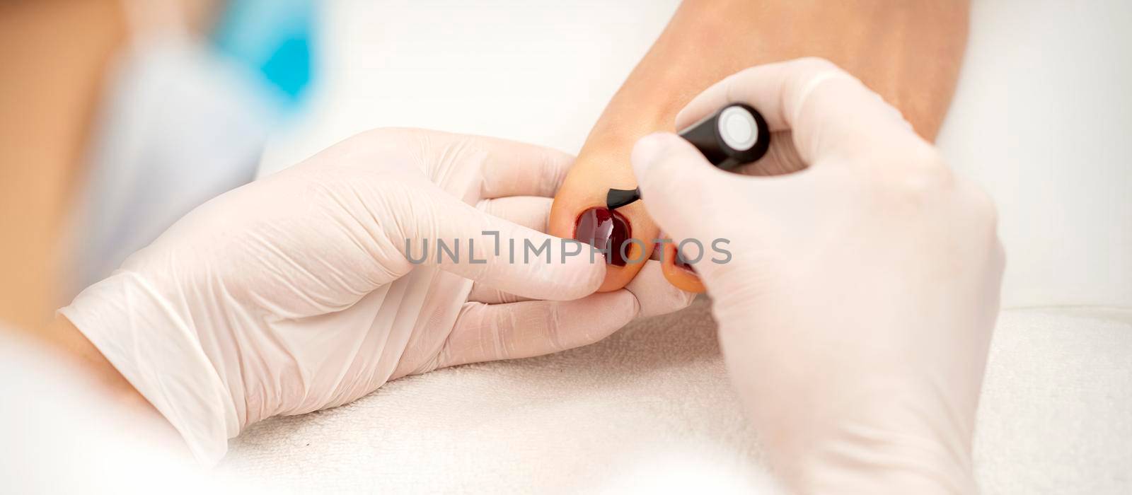 Closeup of hands in white gloves applying burgundy nail polish with a brush on woman's toes in a nail salon