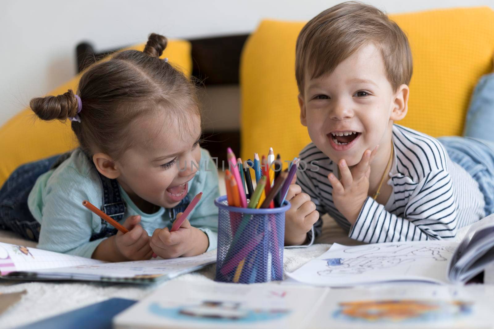smart smiling preshool toddler children boy and girl draw with pencils lying on their stomach at yellow bed. little cute reader have fun, happy kid on quarantine at home. Friendship, family, education.