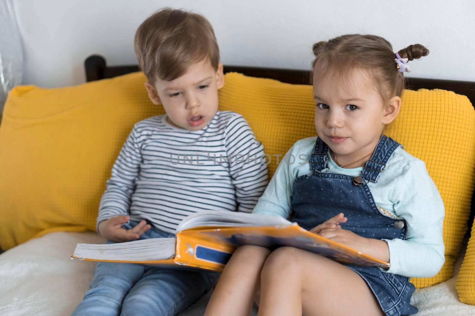 Two preshool, toddler children read large interesting book of fairy tales on yellow bed. Siblings little twins reader have fun, happy kid on quarantine at home. Friendship, family, education concept.