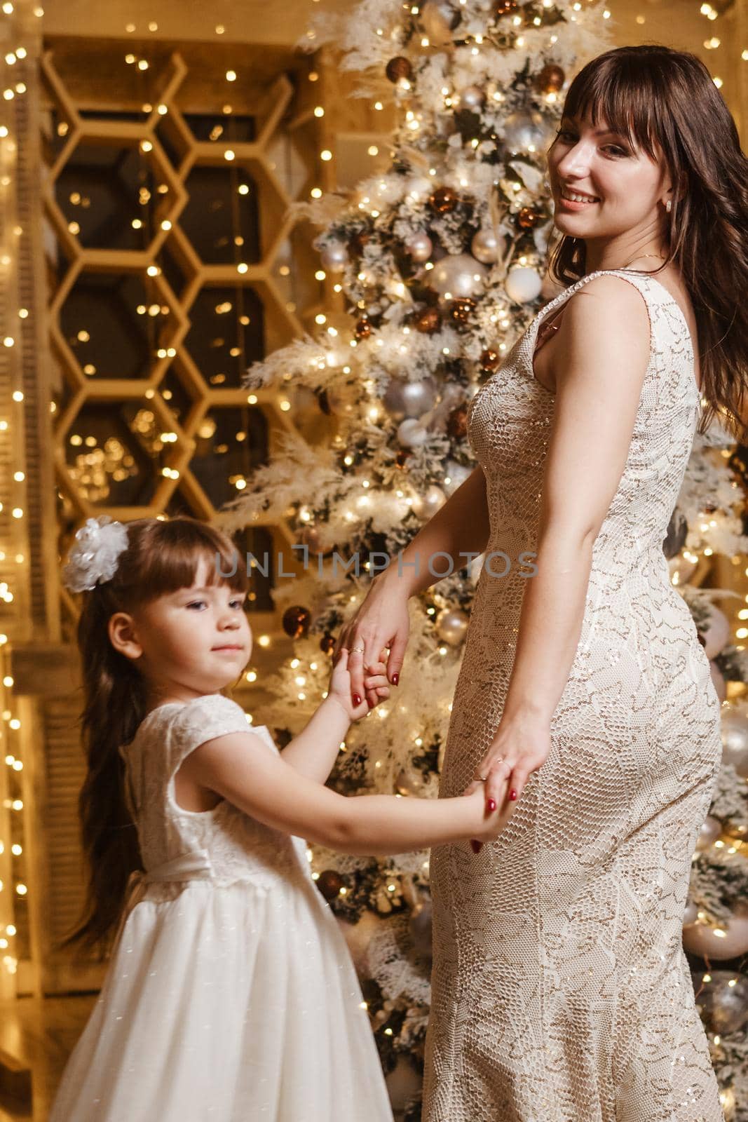 A little girl with her mother in light festive dresses next to the Christmas tree. The theme of New Year's holidays and festive interior with garlands and light bulbs by Annu1tochka