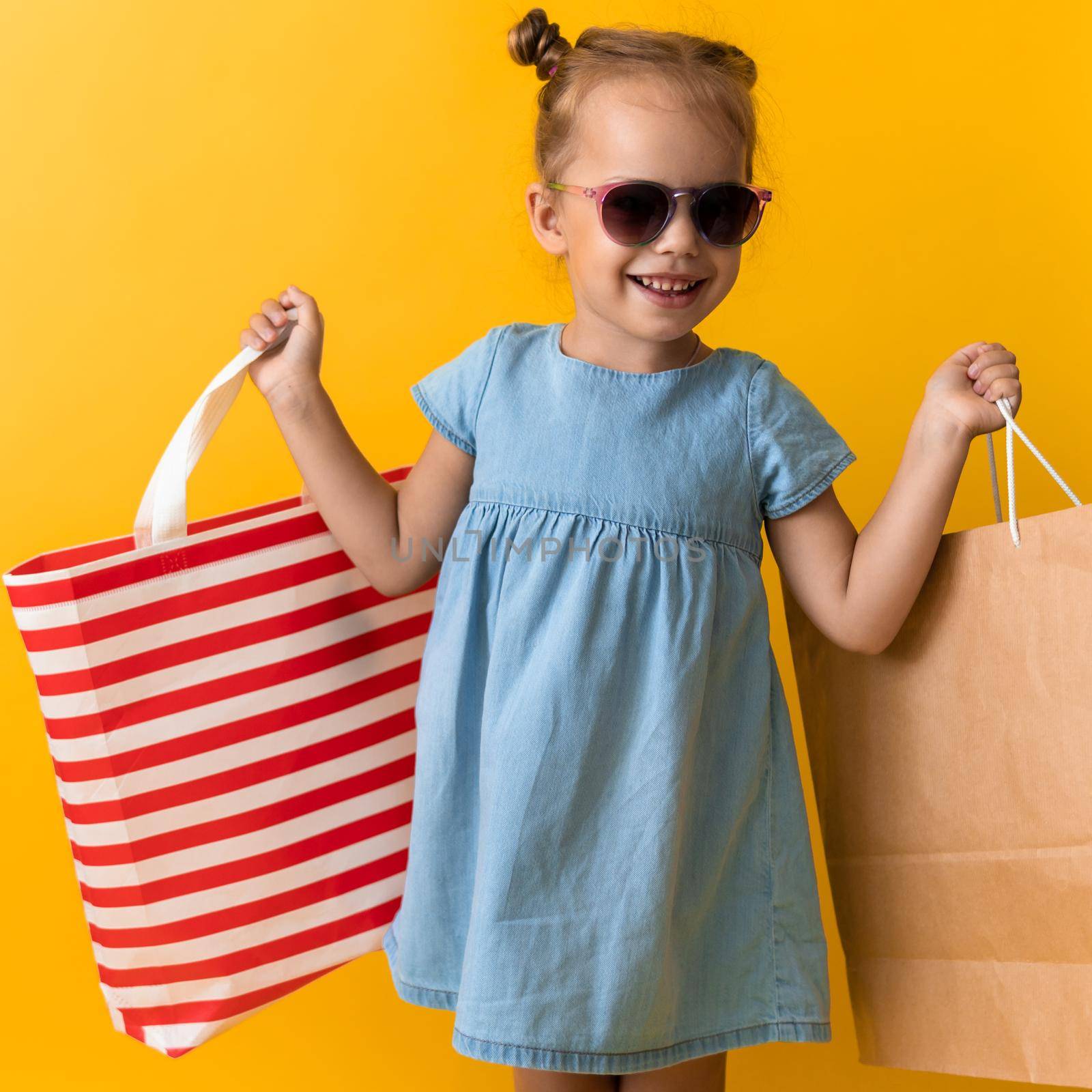 Square Portrait Beautiful Happy Little Preschool Girl In Sunglasses Smiling Cheerful Holding Cardboard Bags Isolated On Orange Yellow background. Happiness, Consumerism, Sale People shopping Concept.