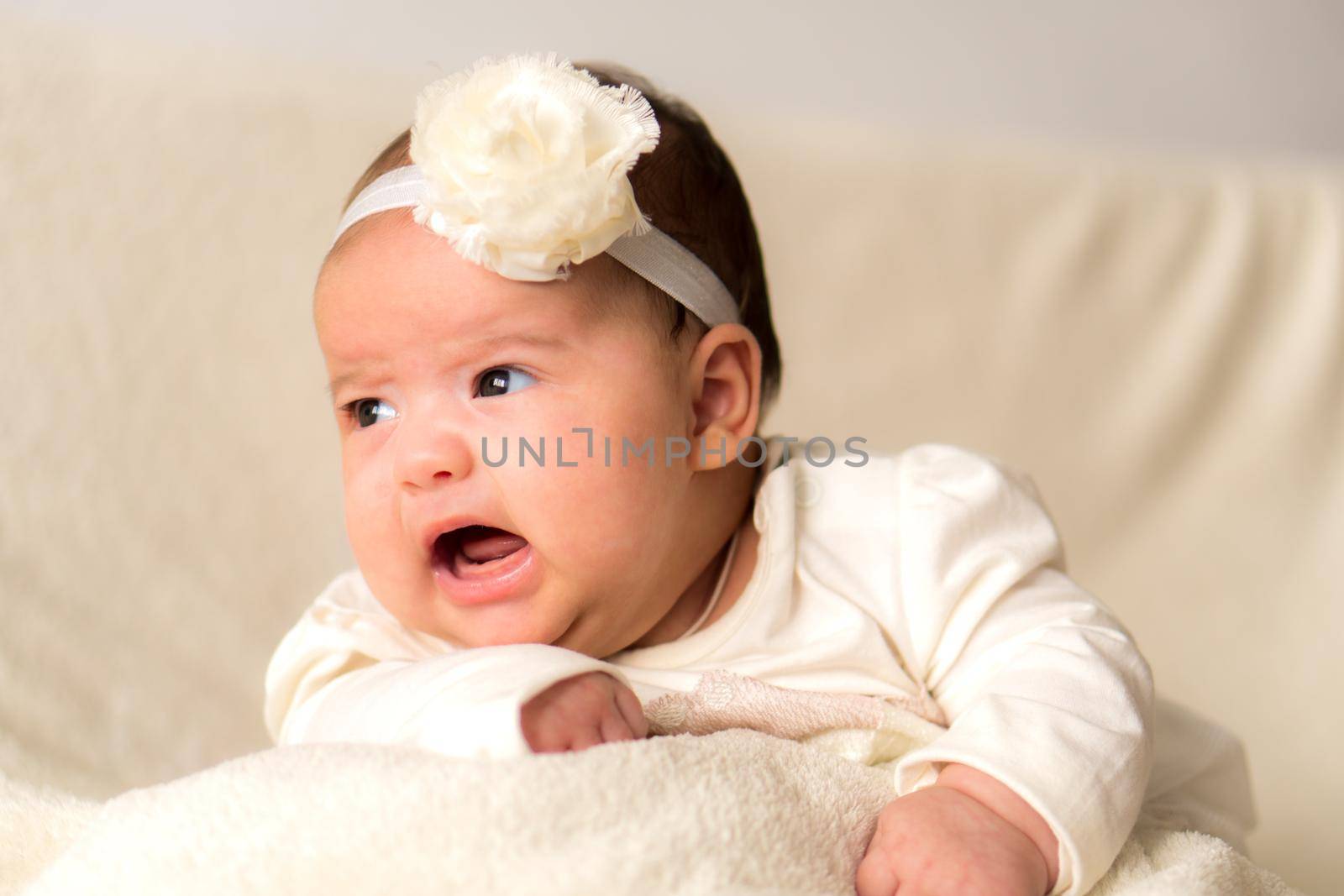 Childhood, motherhood, emotions, fashion concept - worried sad crying chubby baby with open mouth close up little girl in beautiful white dress floral headband lying on soft warm bed on tummy at home.