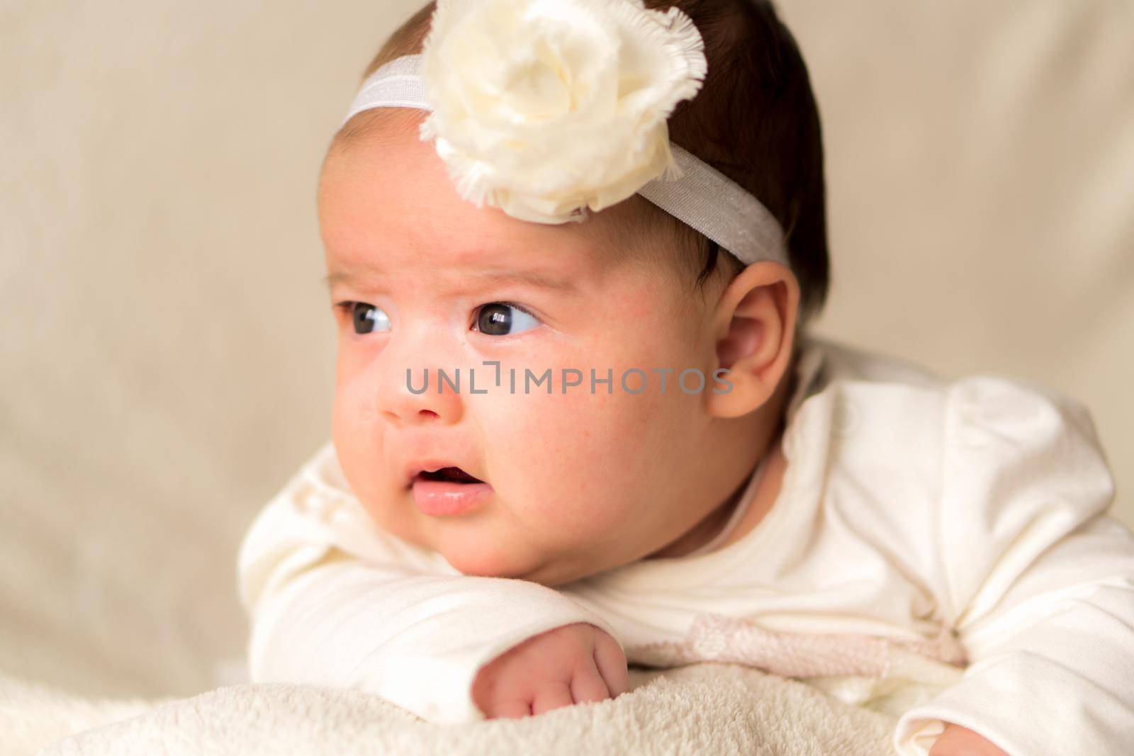 Childhood, motherhood, emotions, fashion concept - worried sad crying chubby baby with open mouth close up little girl in beautiful white dress floral headband lying on soft warm bed on tummy at home.