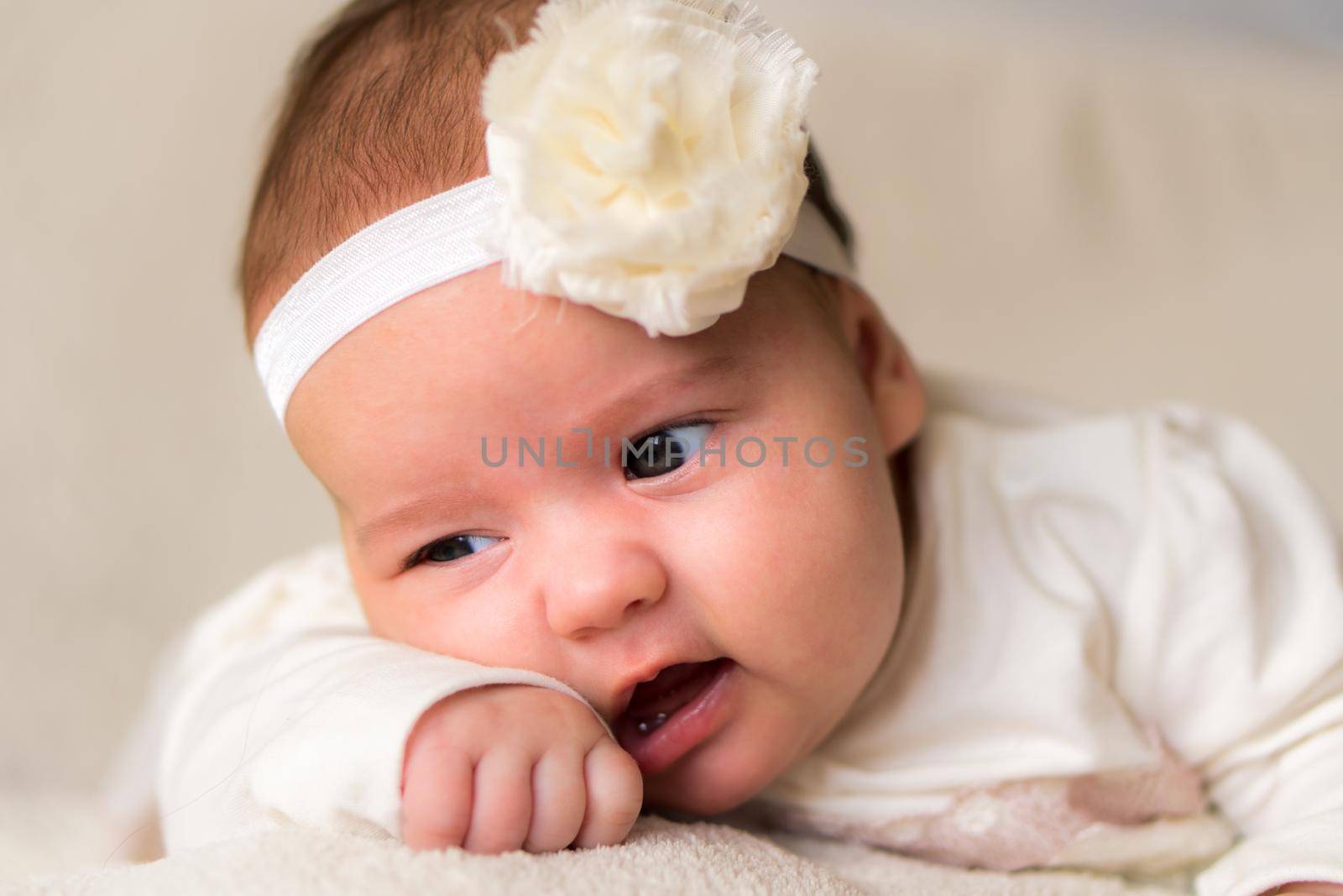 Childhood, motherhood, emotions, fashion concept - worried sad crying chubby baby with open mouth close up little girl in beautiful white dress floral headband lying on soft warm bed on tummy at home.