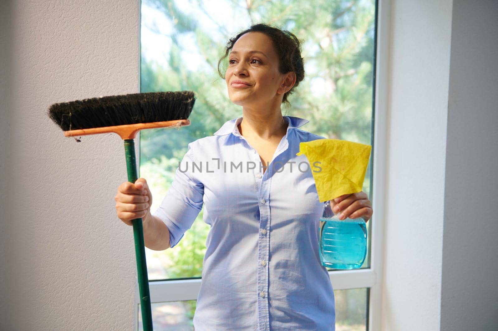 Beautiful Latin American young woman, a pretty housewife cutely smiles looking aside, holding a broom, cloth and detergent while enjoying spring general cleaning in the house. Housekeeping concept