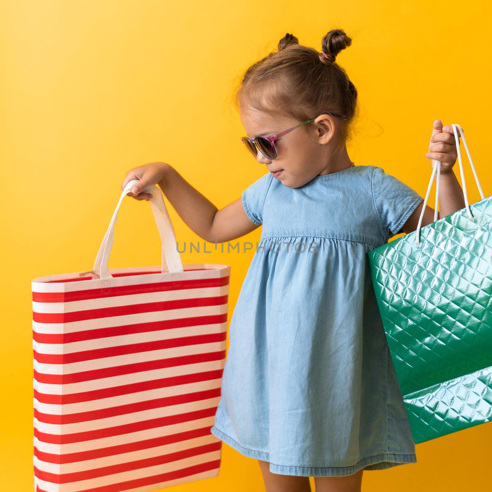 Square Portrait Beautiful Happy Little Preschool Girl In Sunglasses Smiling Cheerful Holding Cardboard Bags Isolated On Orange Yellow background. Happiness, Consumerism, Sale People shopping Concept by mytrykau