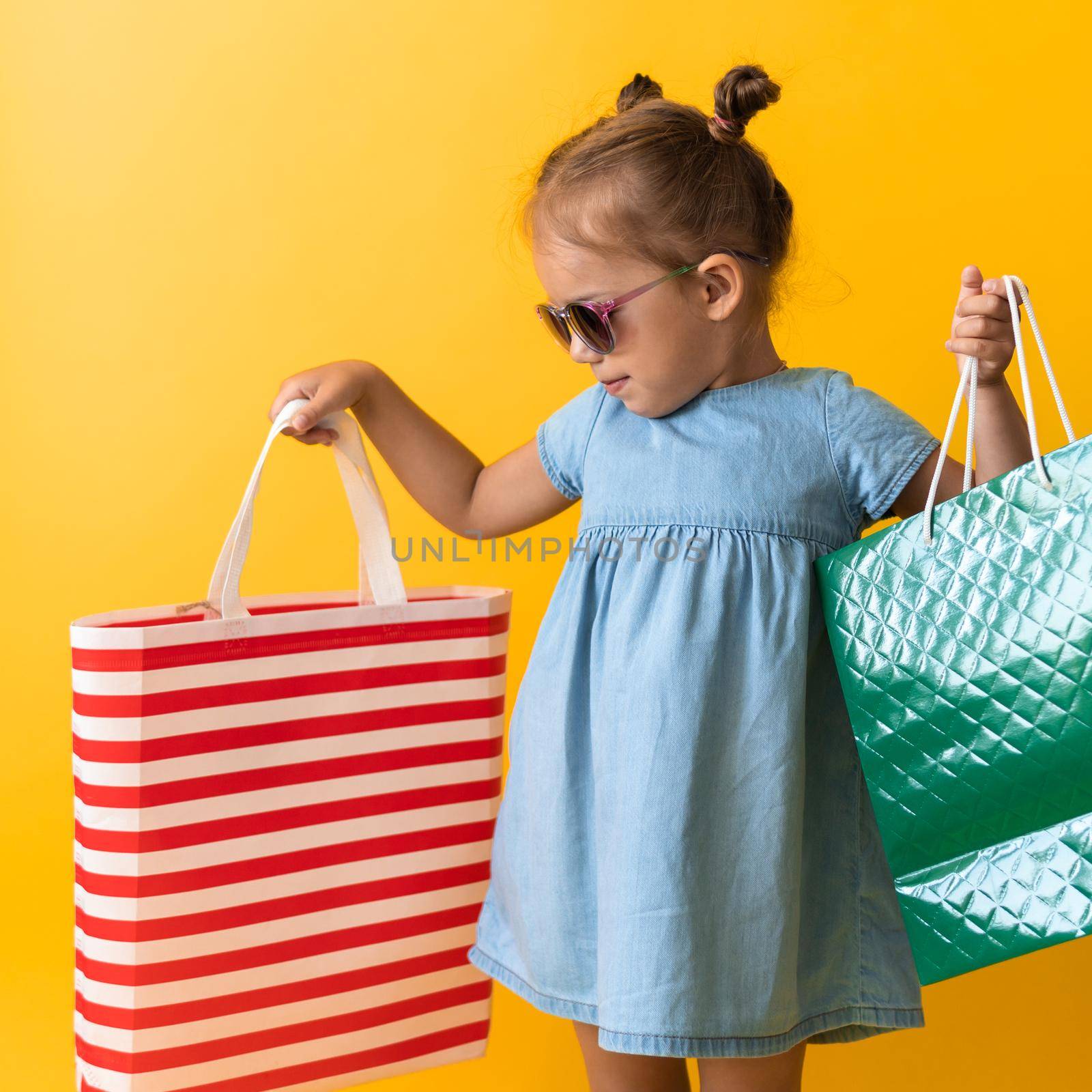 Square Portrait Beautiful Happy Little Preschool Girl In Sunglasses Smiling Cheerful Holding Cardboard Bags Isolated On Orange Yellow background. Happiness, Consumerism, Sale People shopping Concept.