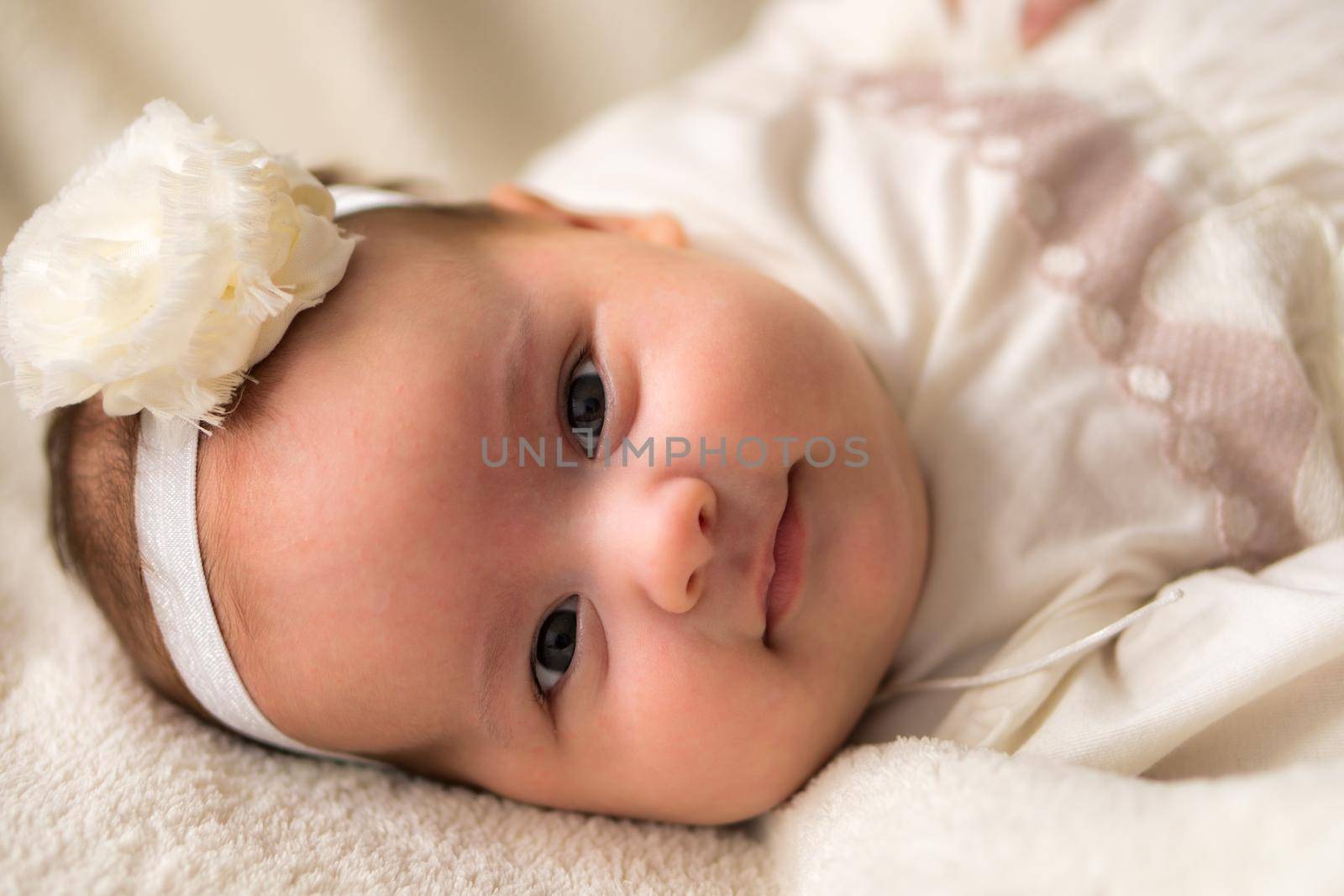 Childhood, motherhood, emotions, fashion concepts - Cute smiling happy chubby baby close up little girl in beautiful white dress flower headband on head lying on soft warm bed on her tummy at home.
