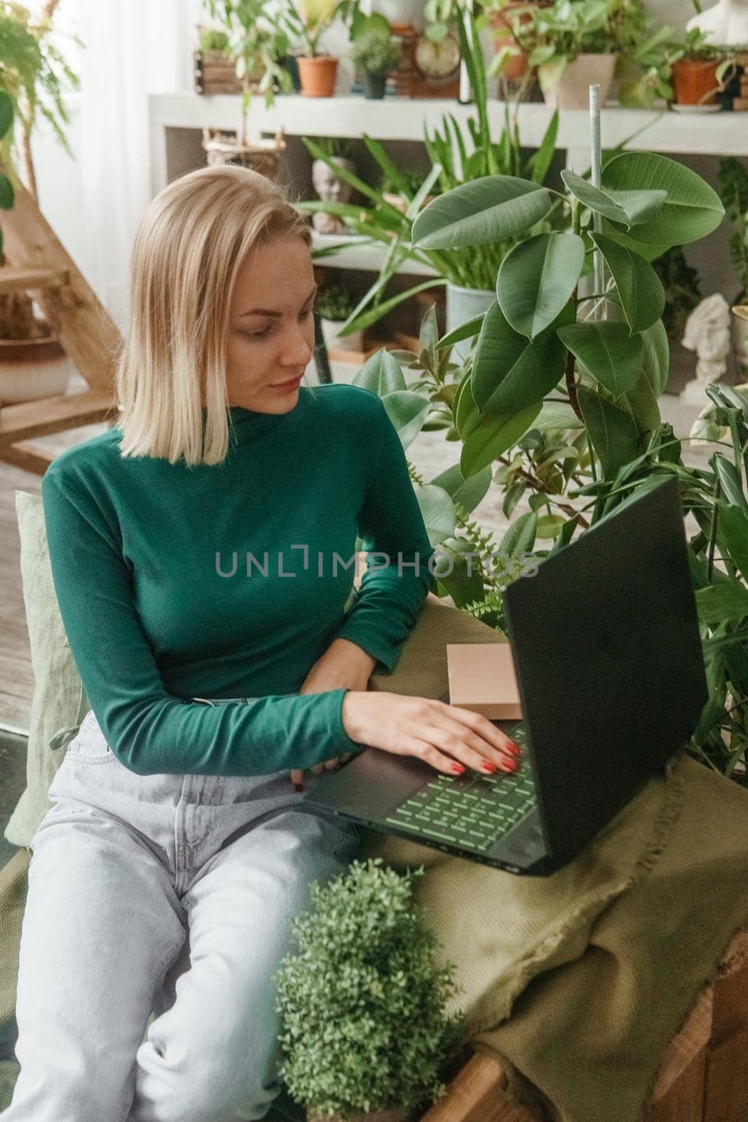 A blonde woman in a room with a lot of green indoor plants is working on a laptop. The concept of biophysical design in the interior. Work from home, work as a freelancer.