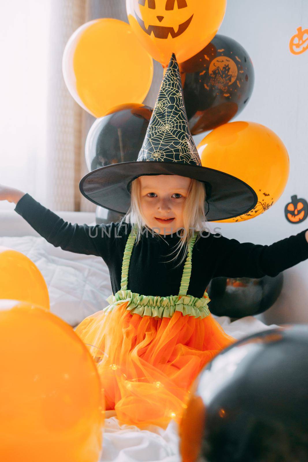 Children's Halloween - a girl in a witch hat and a carnival costume with airy orange and black balloons at home. Ready to celebrate Halloween.