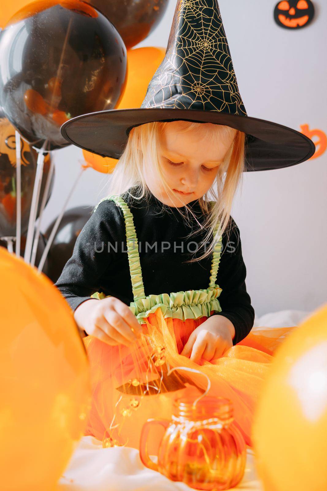 Children's Halloween - a girl in a witch hat and a carnival costume with airy orange and black balloons at home. Ready to celebrate Halloween.