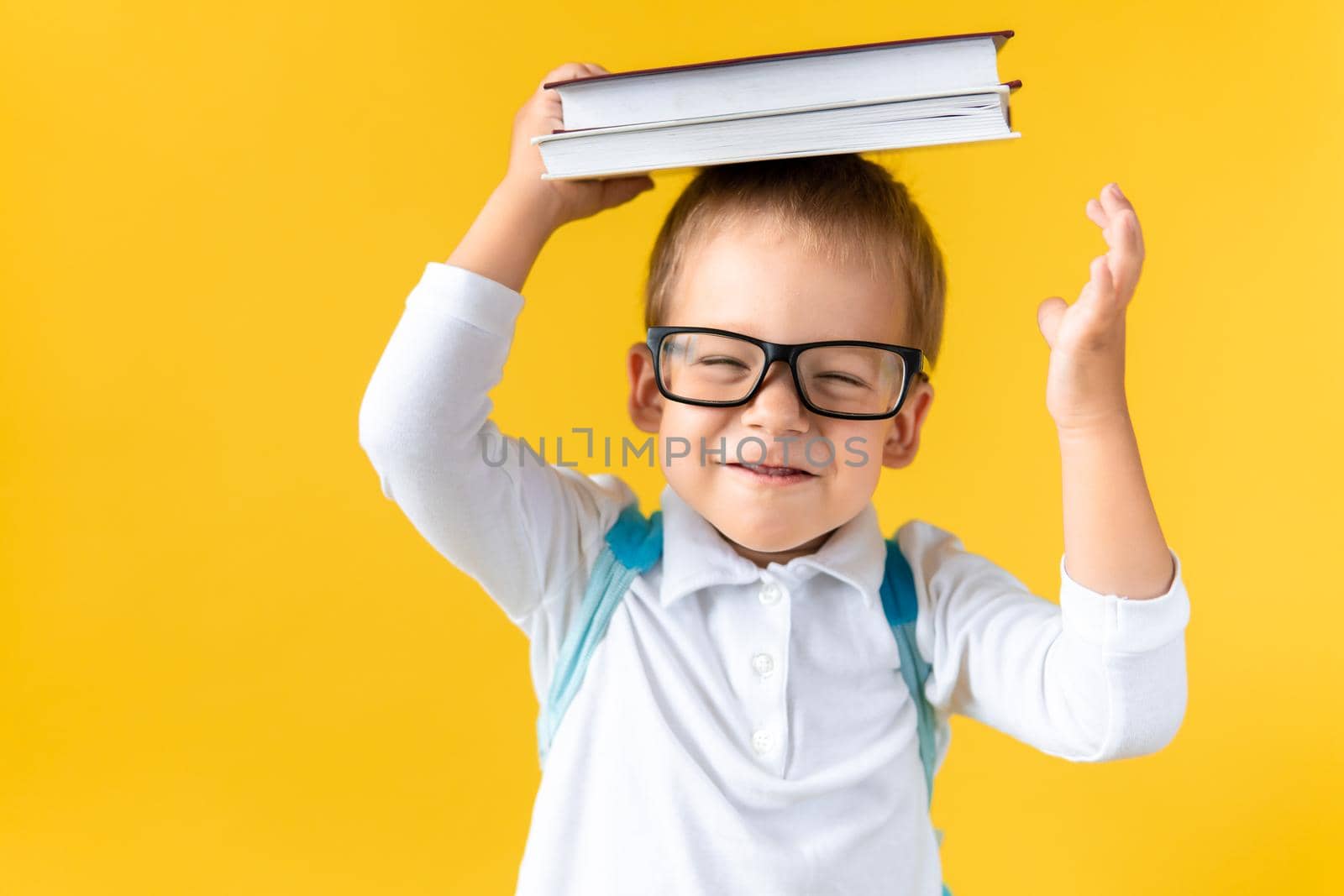 Funny Preschool Child Boy in Glasses with Book on Head and Bag on Yellow Background Copy Space. Happy Smiling Kid Go Back to School, Kindergarten. Success, Motivation, Winner Genius, Superhero concept.